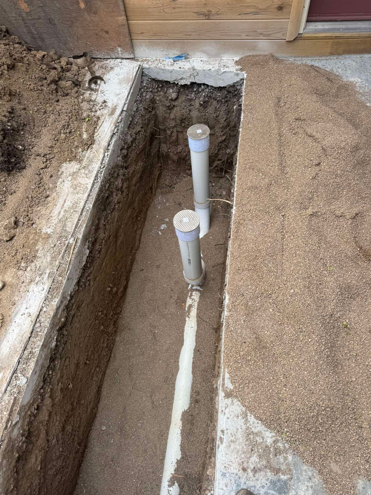 A rectangular trench dug in the ground featuring two vertical white PVC pipes connected by a horizontal base pipe.