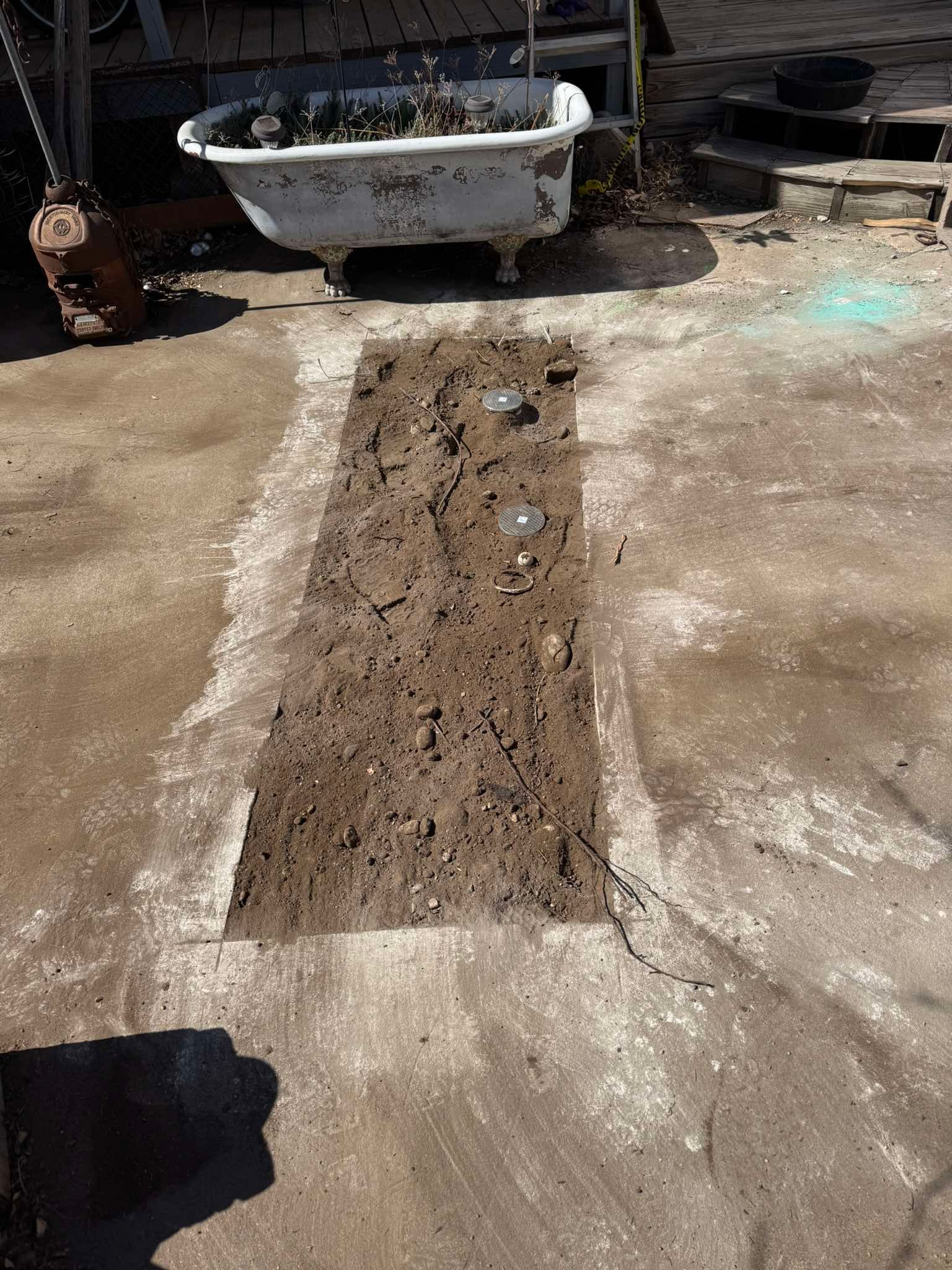 A rectangular section of concrete has been removed from a patio, exposing the dirt beneath, near a vintage bathtub planter.