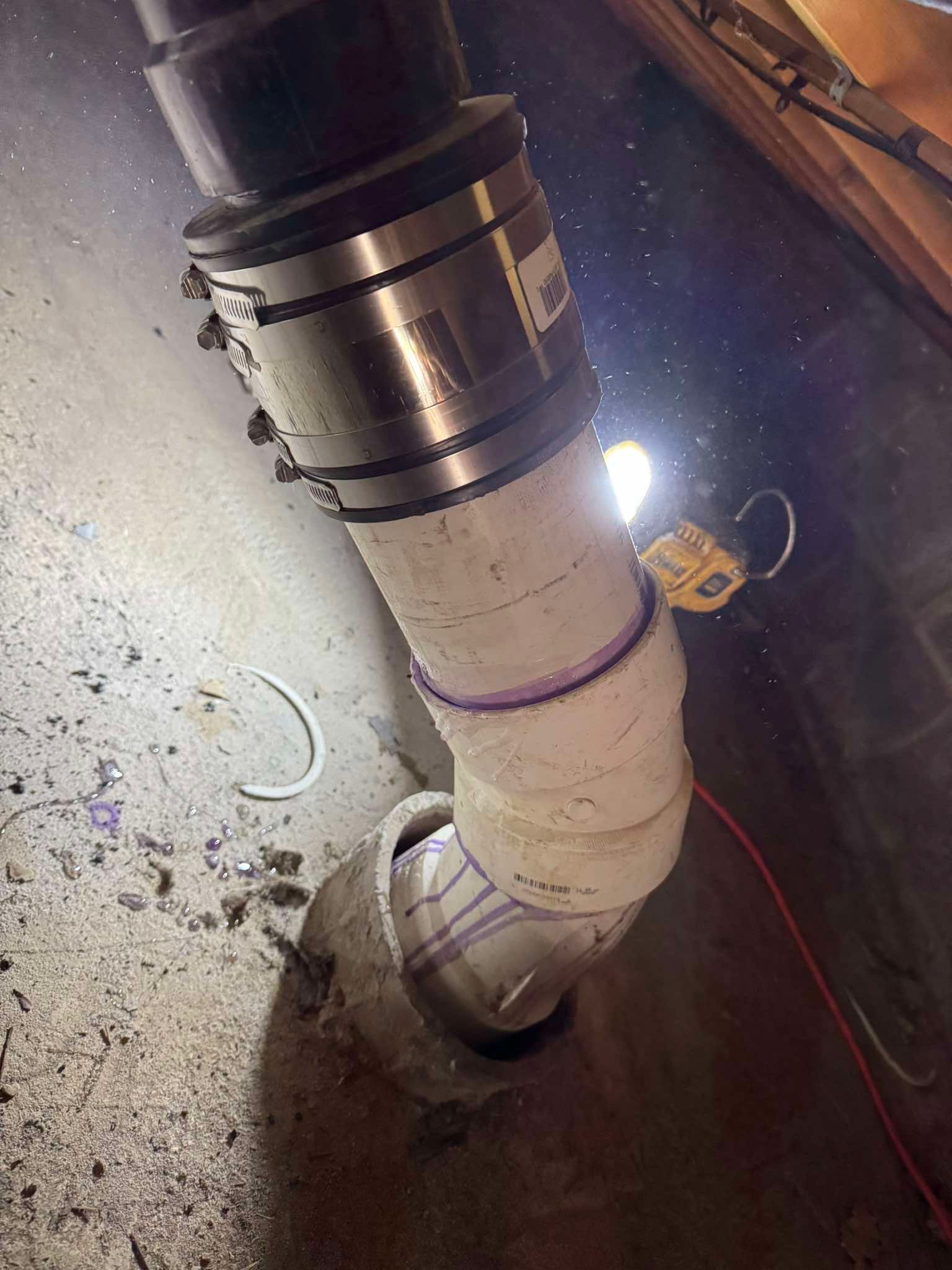 A white PVC pipe connected to a larger black pipe via a metal shielded coupling, emerging from a concrete floor.