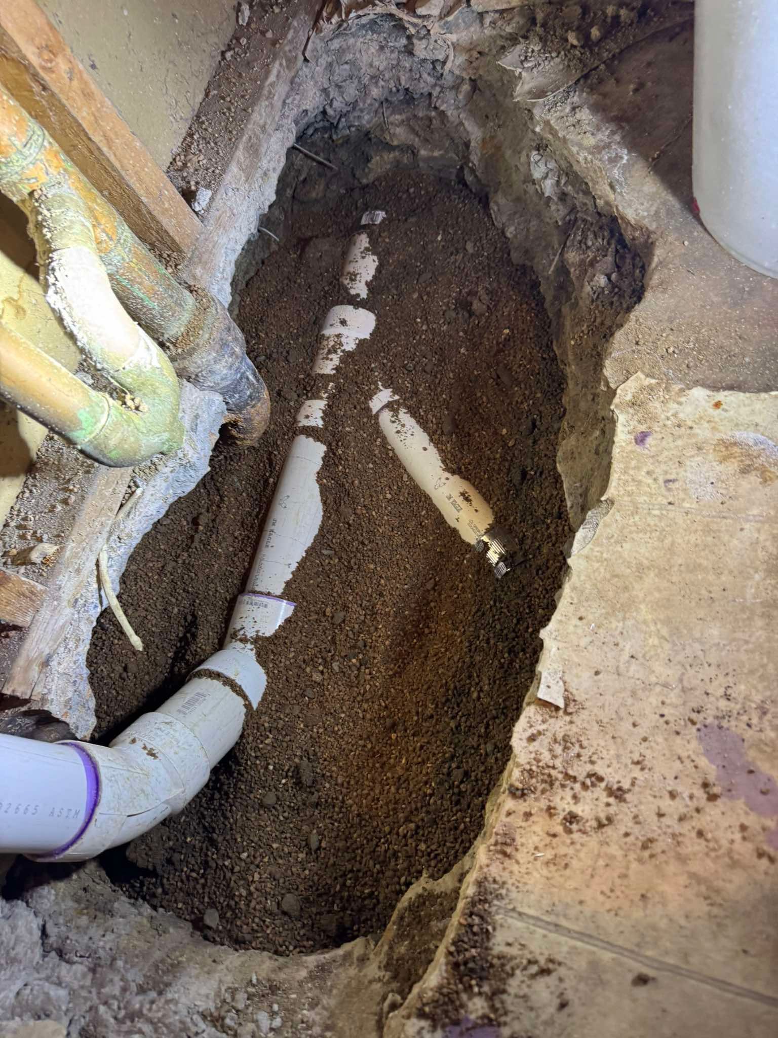 A section of white PVC plumbing pipes exposed in a dirt-filled trench cut through a concrete floor.