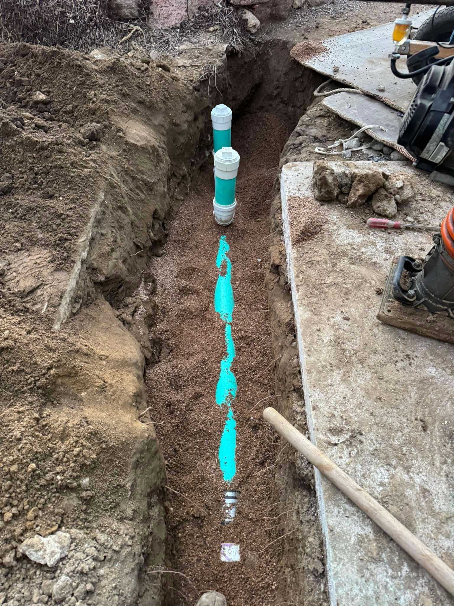 A trench containing a green pipe with vertical capped risers, next to a concrete slab and dirt banks.