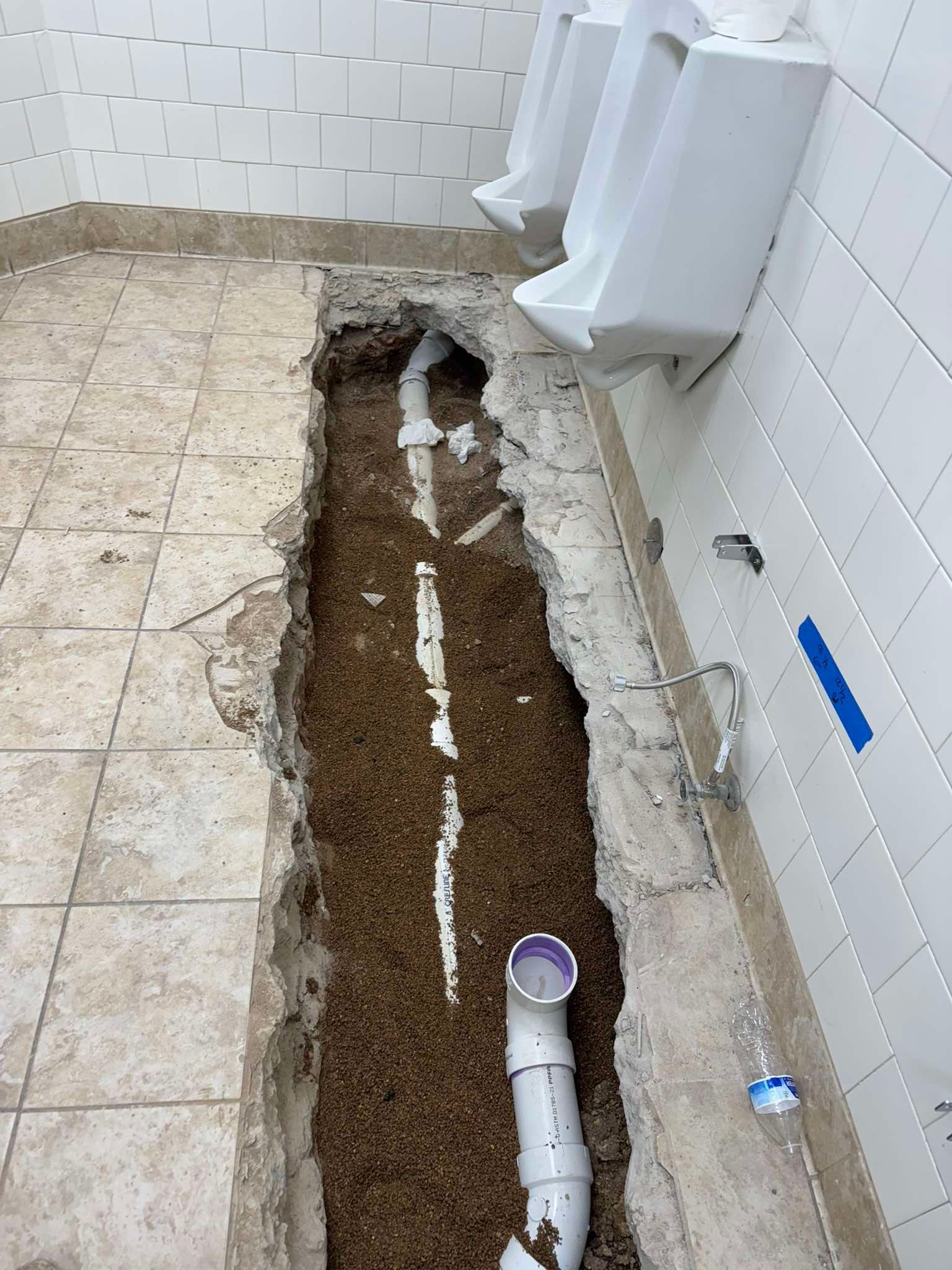 A restroom with a trench cut into the floor, revealing white plumbing pipes beneath wall-mounted urinals.