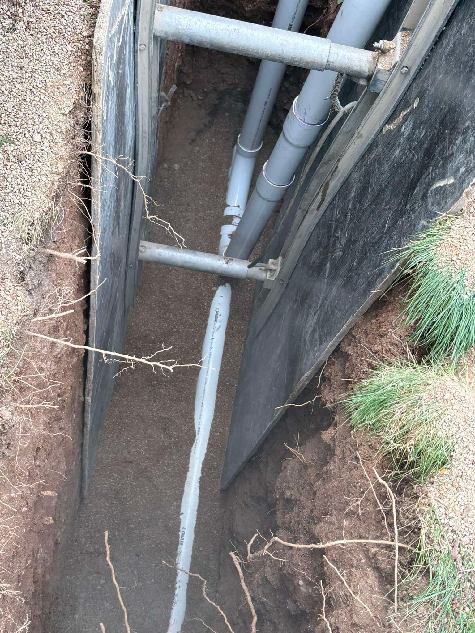 Gray plastic pipes run through a narrow, shored dirt trench in the ground.