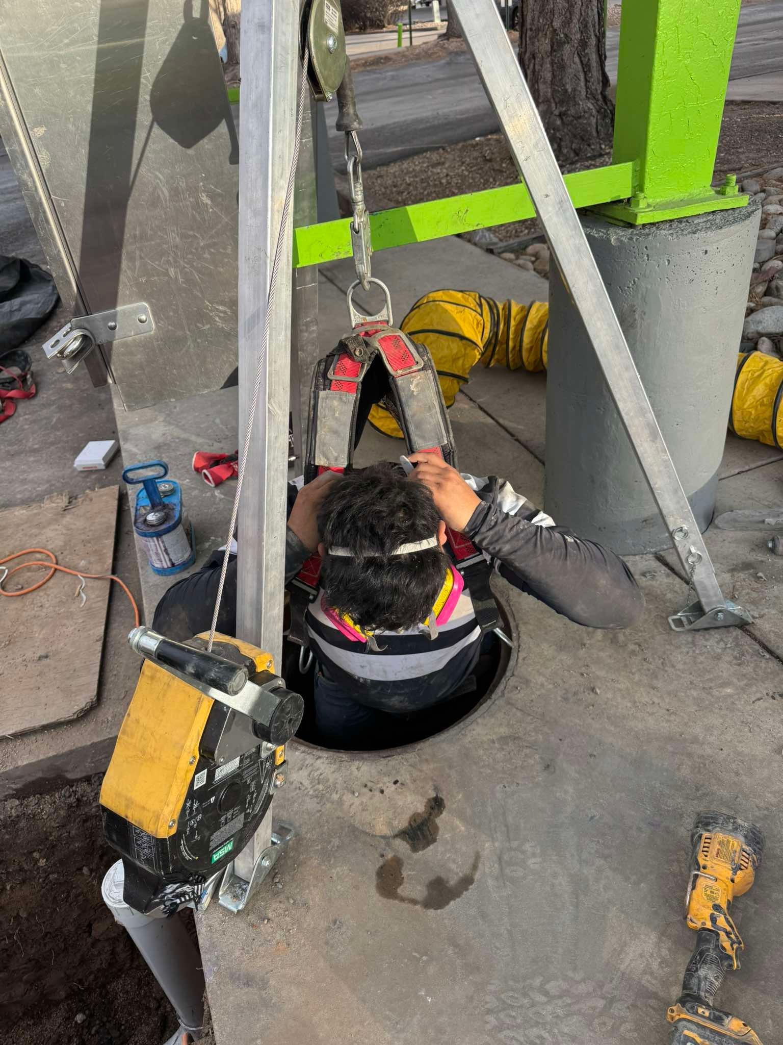 A worker in safety gear enters an underground space through a manhole, secured by a tripod winch system on a sidewalk.