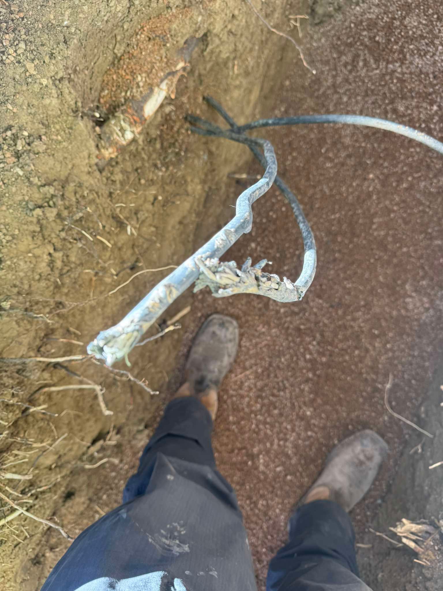 A person’s legs and feet wearing boots stand near a damaged underground cable exposed in a trench.