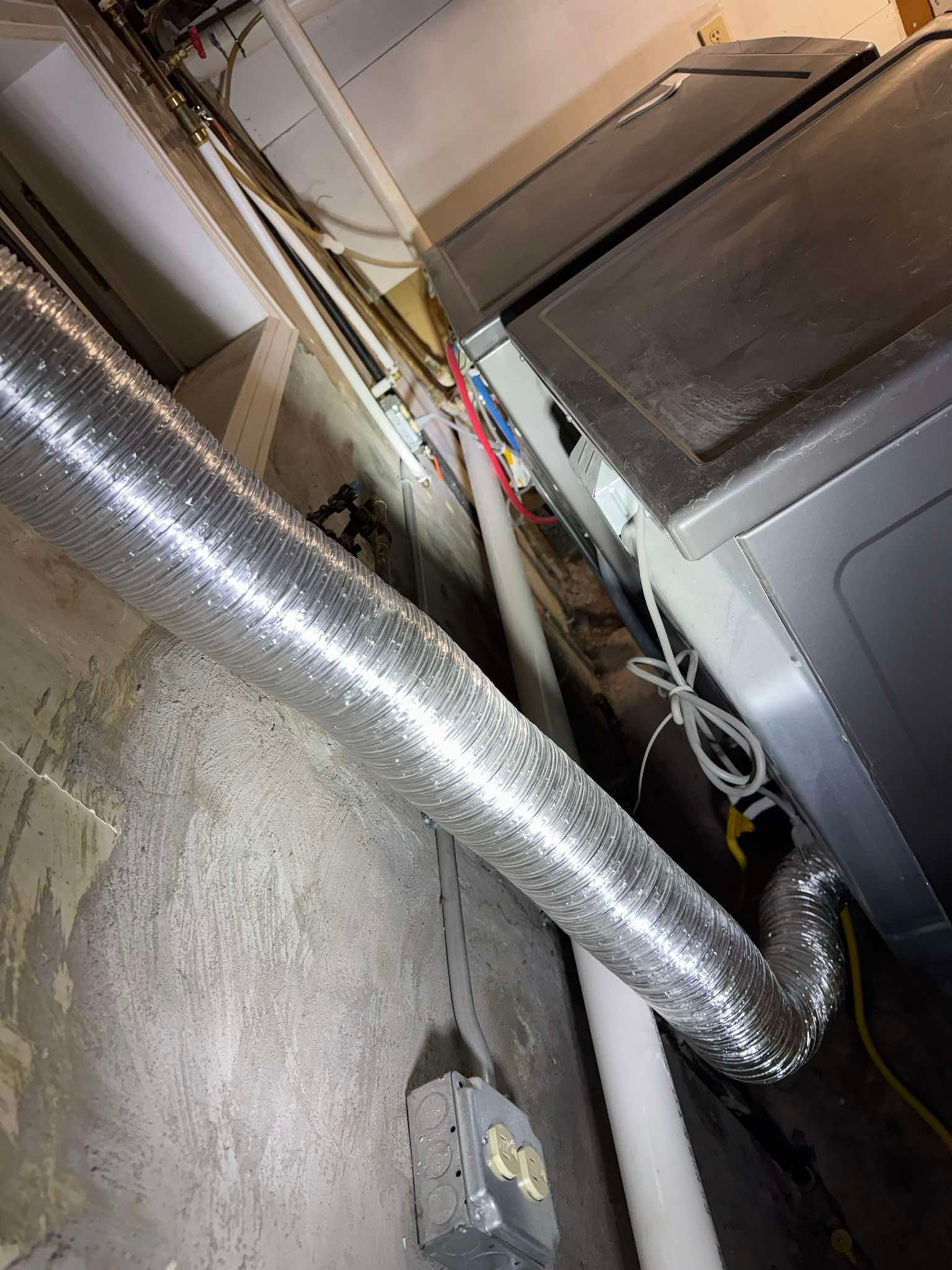 A metallic dryer vent hose runs across a concrete basement floor next to a dark-colored appliance and white pipes.