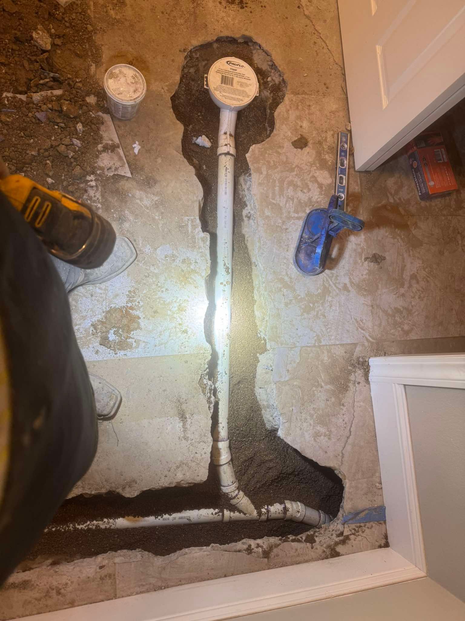 White PVC plumbing pipes installed in a narrow trench cut into a concrete floor, with a cleanout cap and tools nearby.