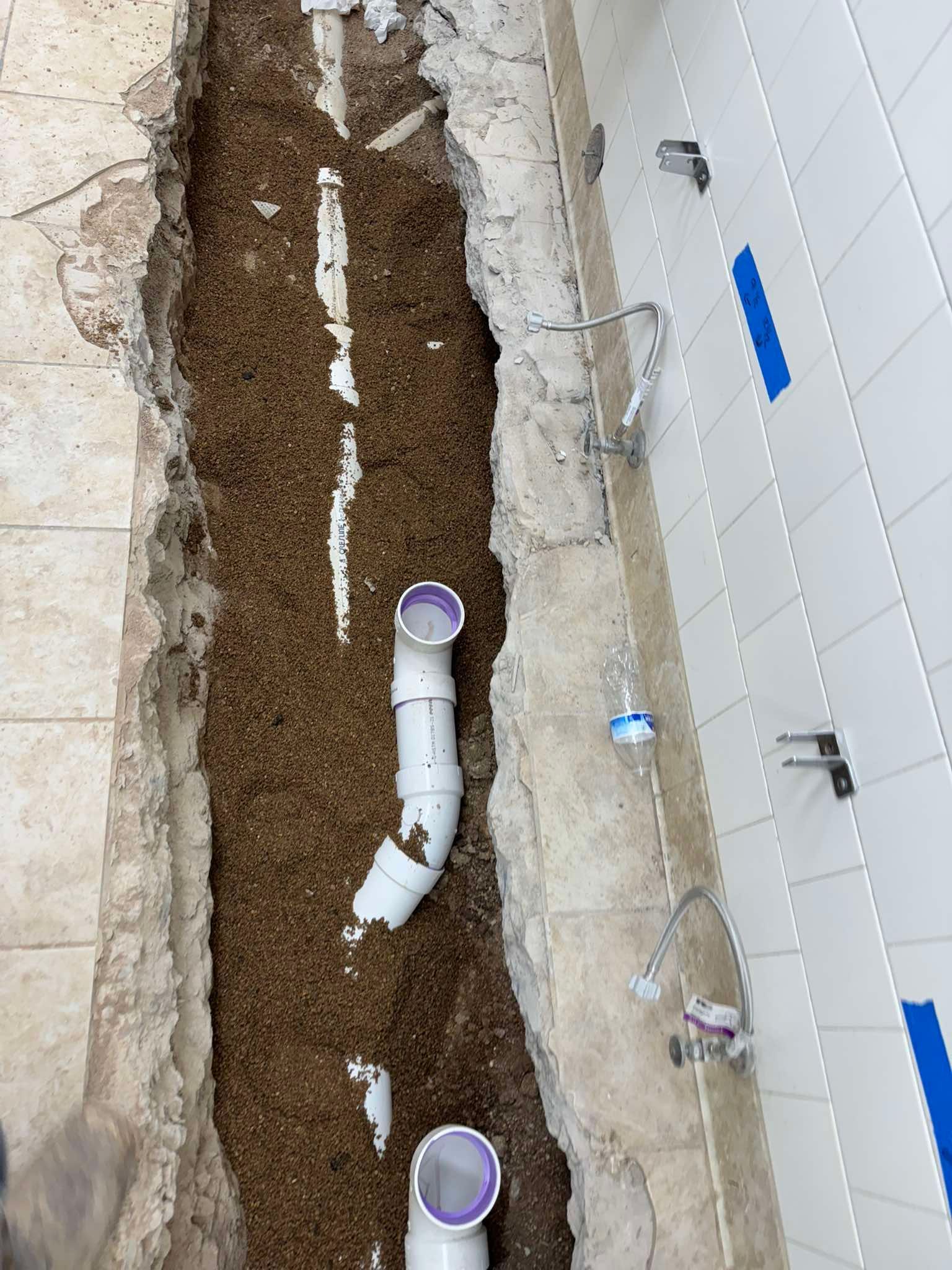 A construction trench in a tiled floor showing exposed white PVC plumbing pipes connected to two upward-facing drains.
