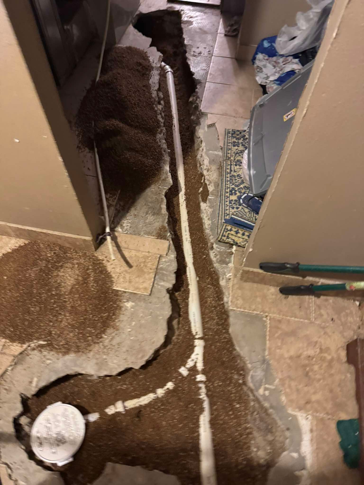 A trench cut through a concrete floor to expose white PVC plumbing pipes connected to a cleanout drain.