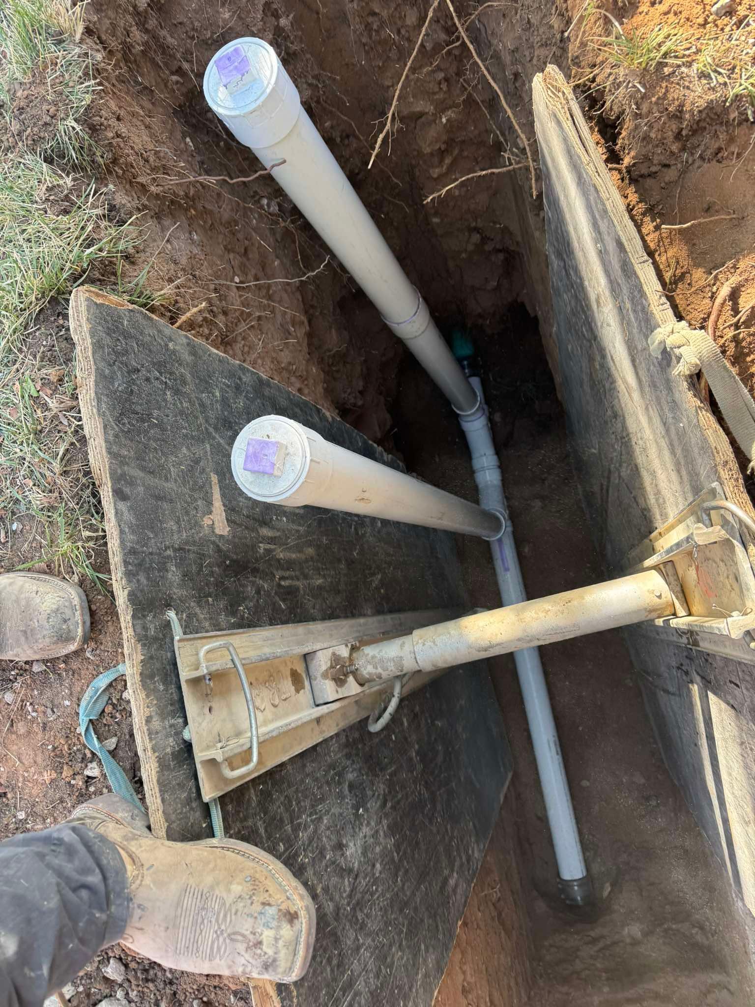 A trench with a metal safety shoring box, exposed PVC pipes, and a worker's boot visible in the bottom left corner.