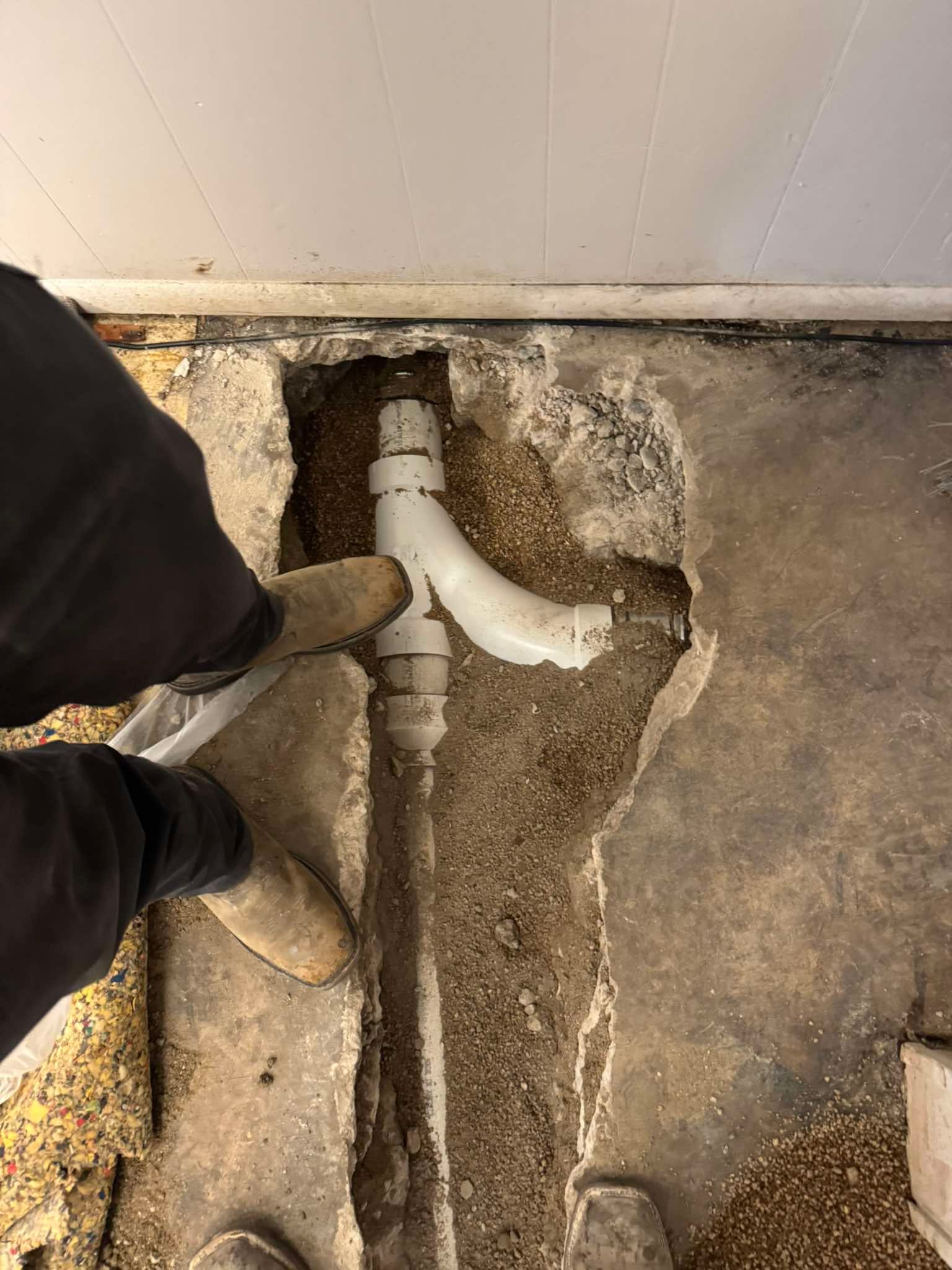 A person’s feet stand on a concrete floor next to an exposed white PVC drain pipe with a Y-fitting in a trench.