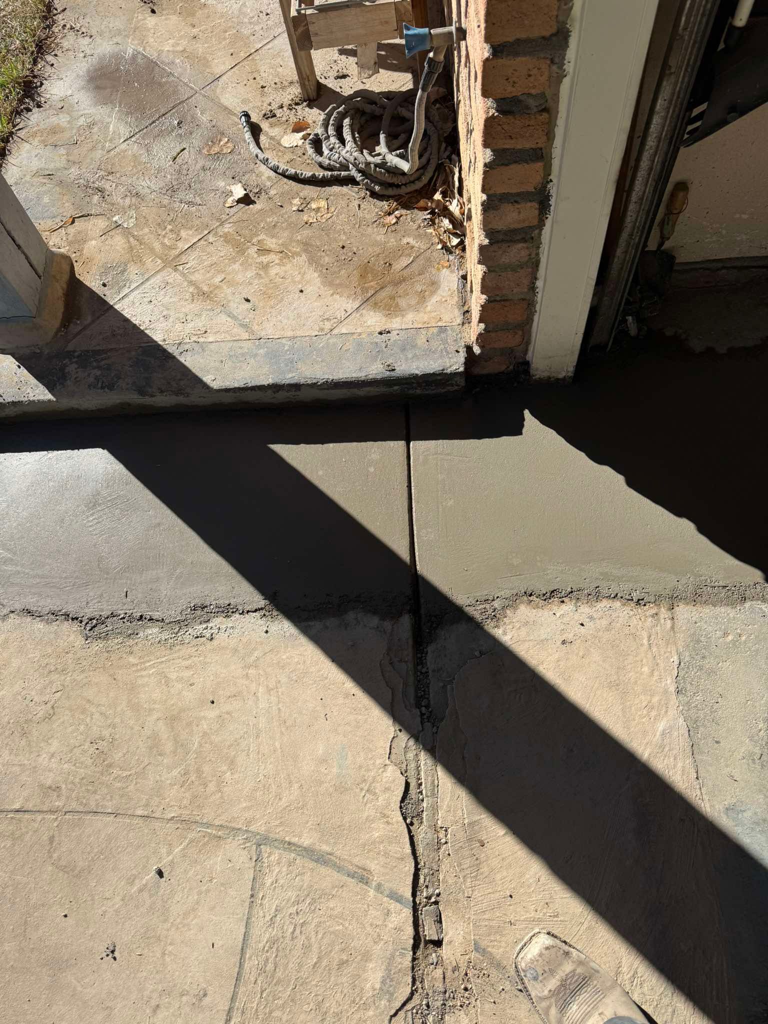 A section of new concrete poured against an older, cracked concrete walkway near a brick wall and a doorway.