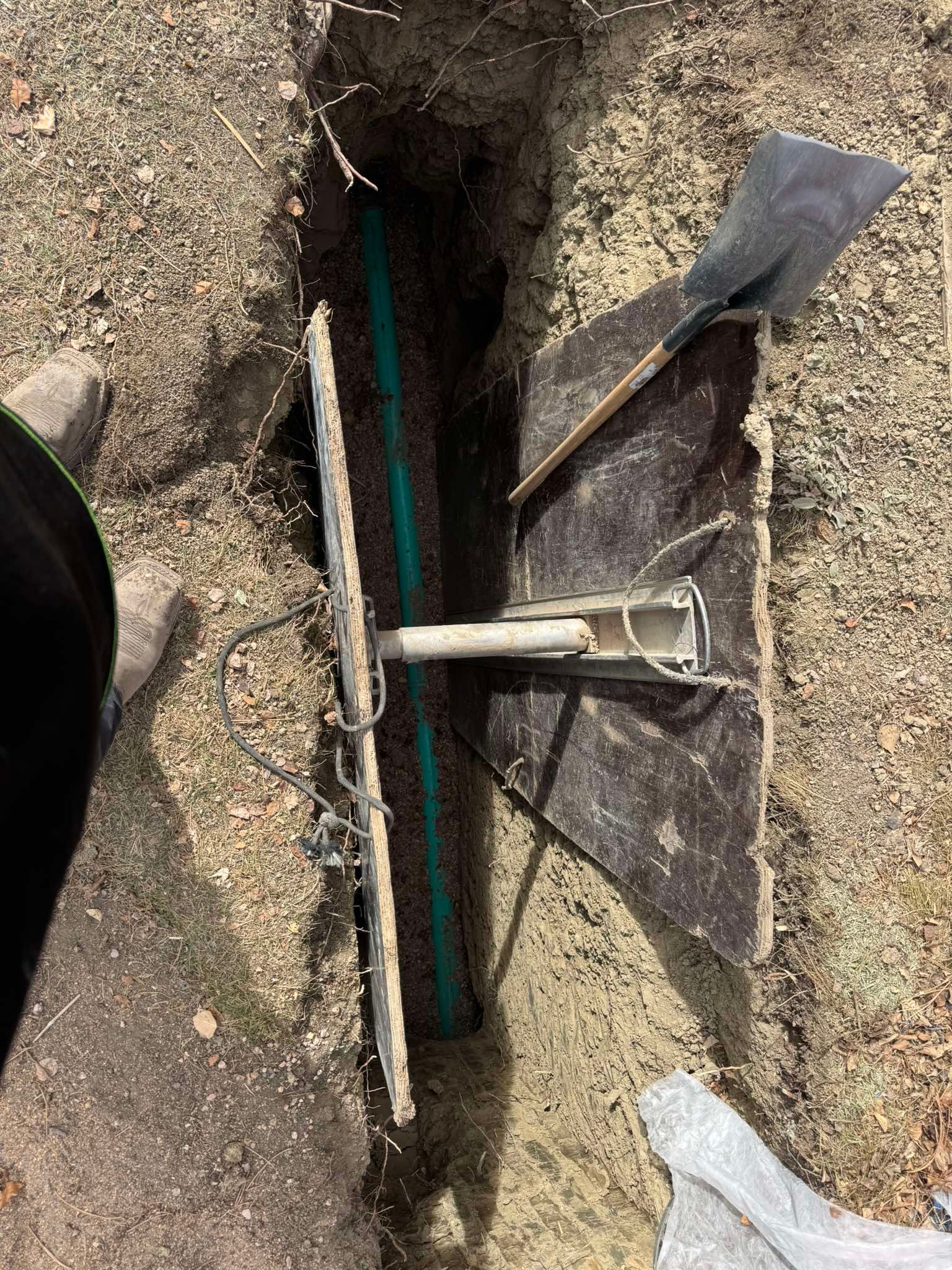A trench with a green pipe at the bottom, covered by wooden boards with metal brackets, next to a shovel on the ground.