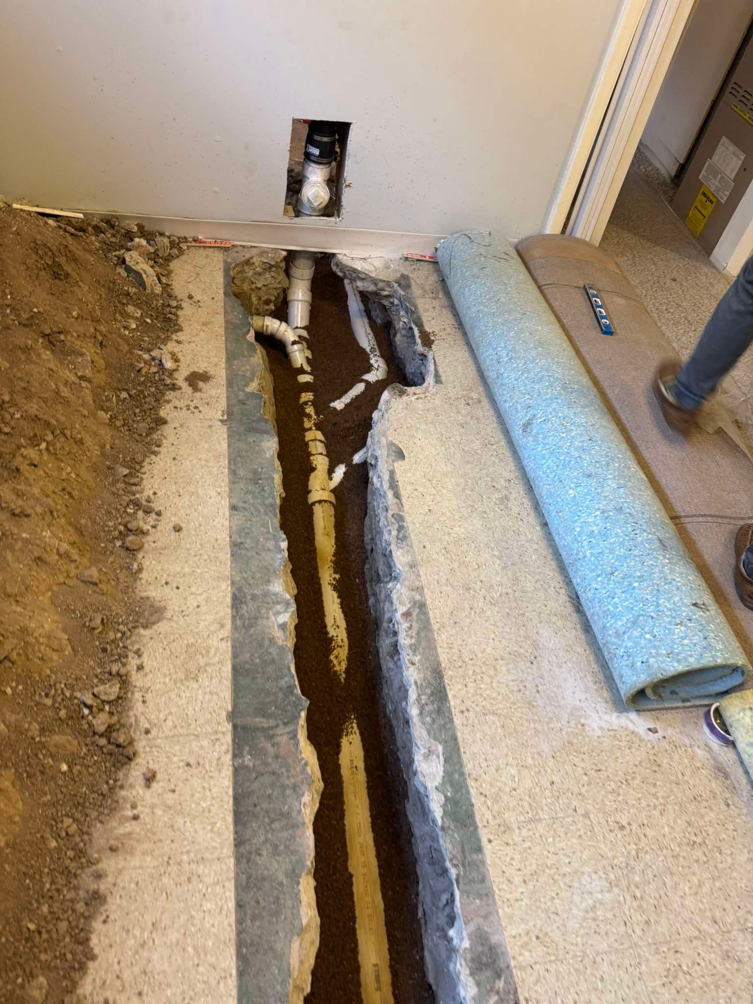 Concrete floor cut open to expose pipes and soil in a room with rolled-up flooring materials.