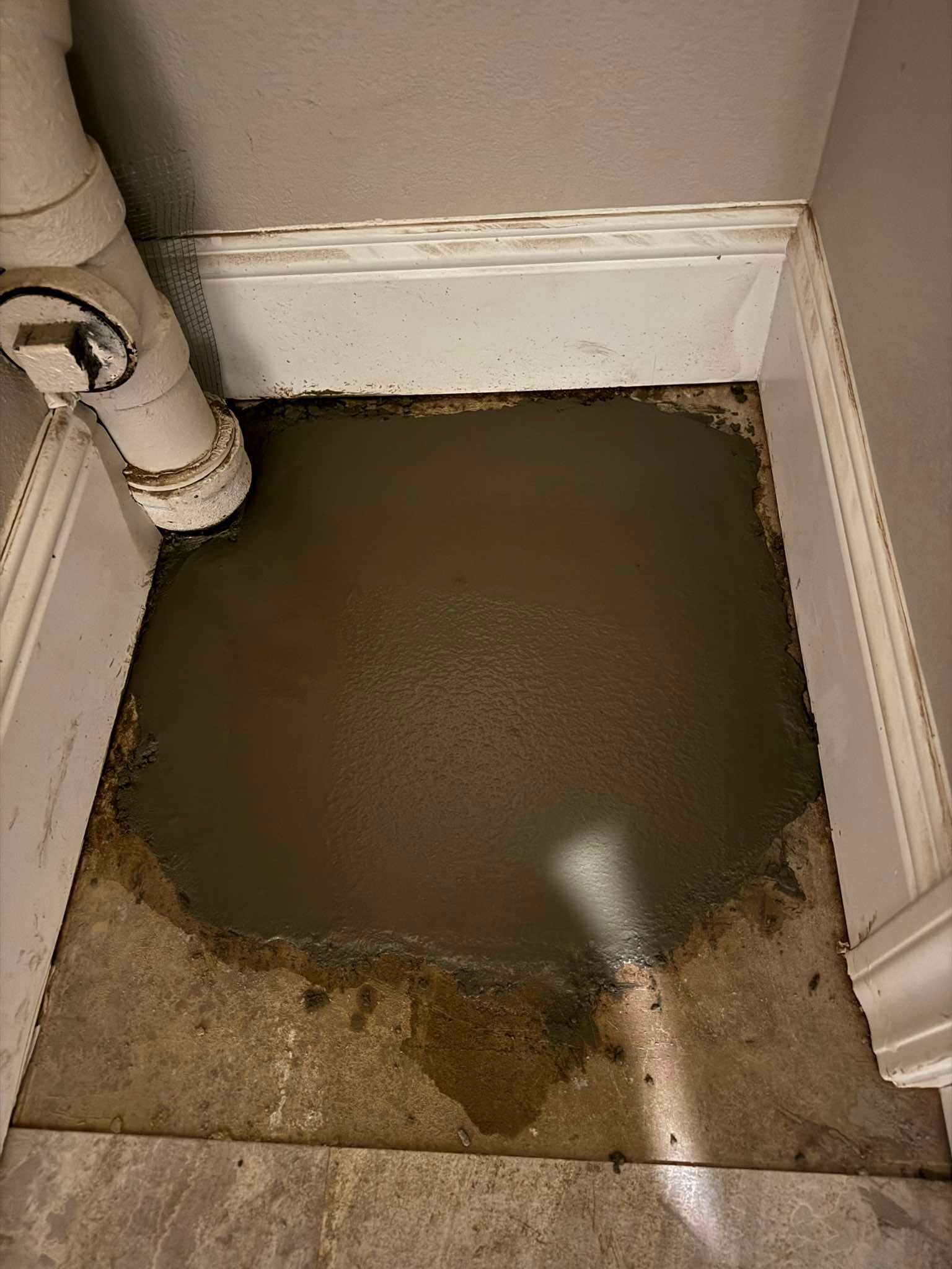 A patch of wet, gray cement on a floor next to a white PVC pipe in a corner against white baseboards.
