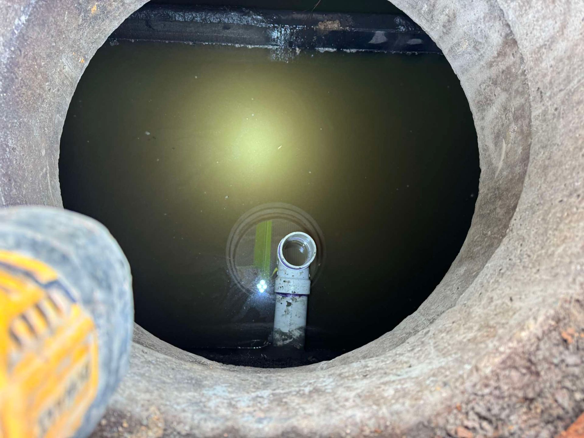 A view into a dark, water-filled utility pit featuring a white PVC pipe elbow sticking up from the murky water.