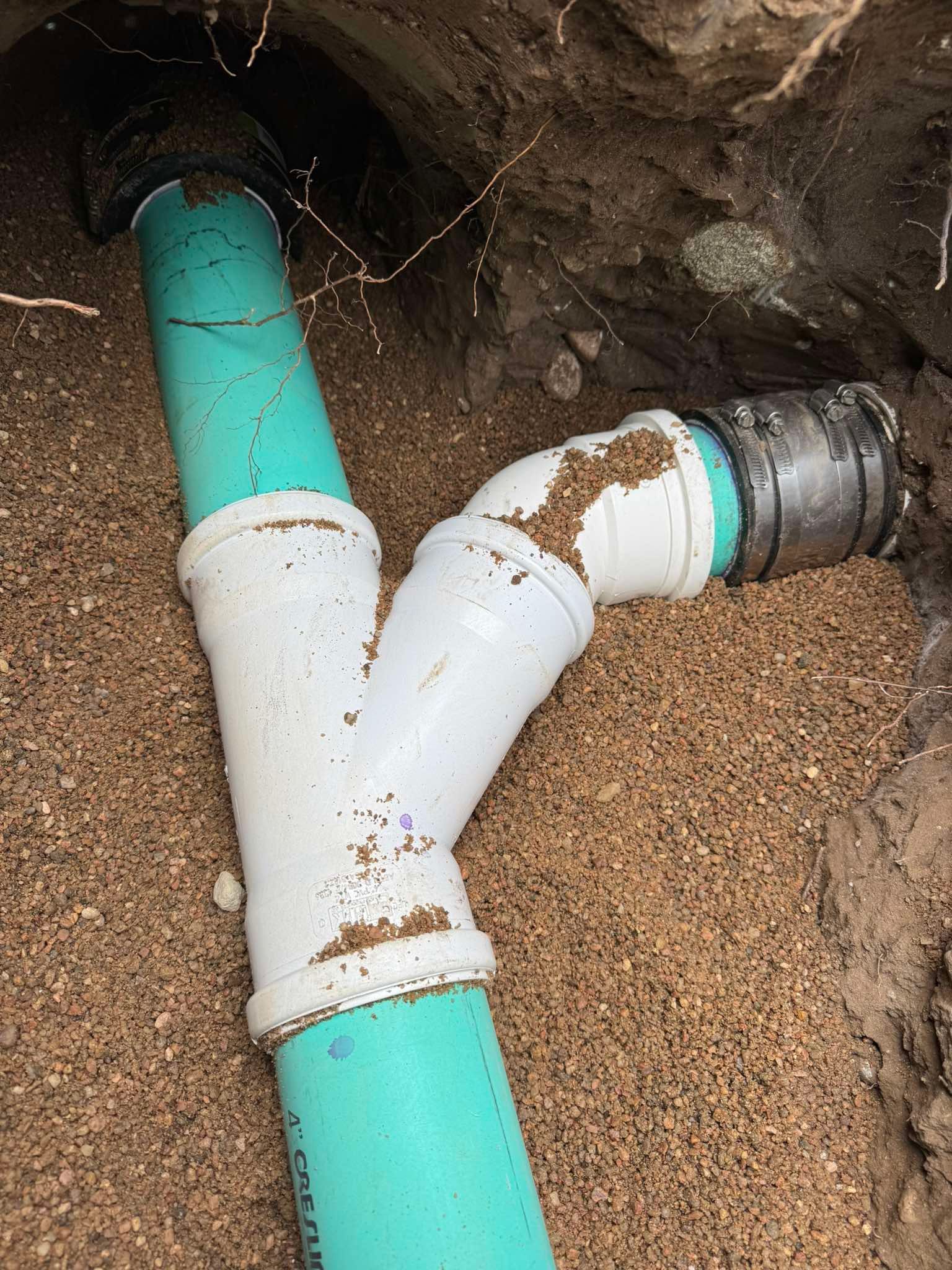 A white PVC wye fitting connects green pipes in a trench filled with gravel, including a mechanical rubber coupling.