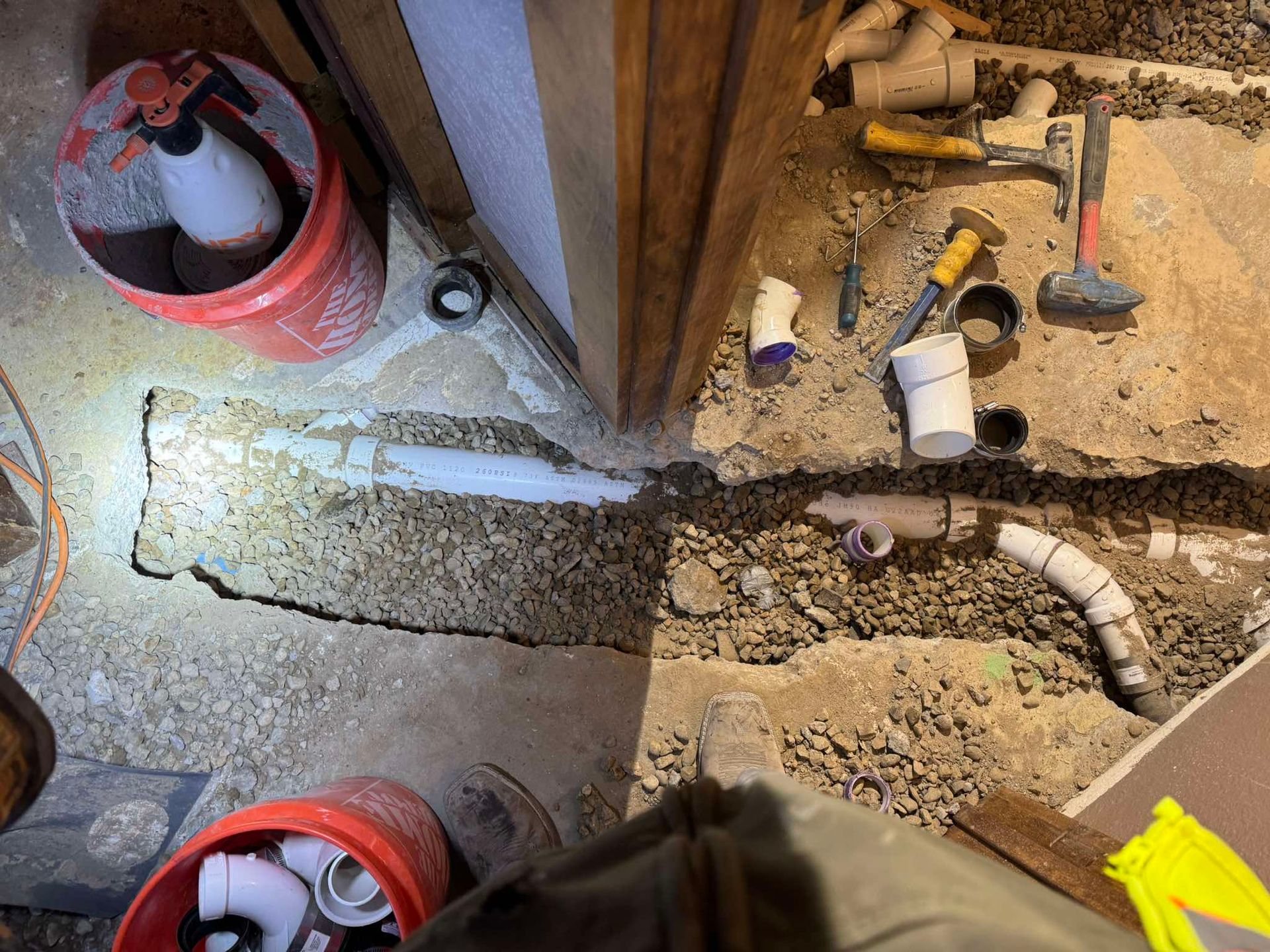 A plumbing repair site on a concrete floor with exposed white pipes, assorted tools, and red buckets.