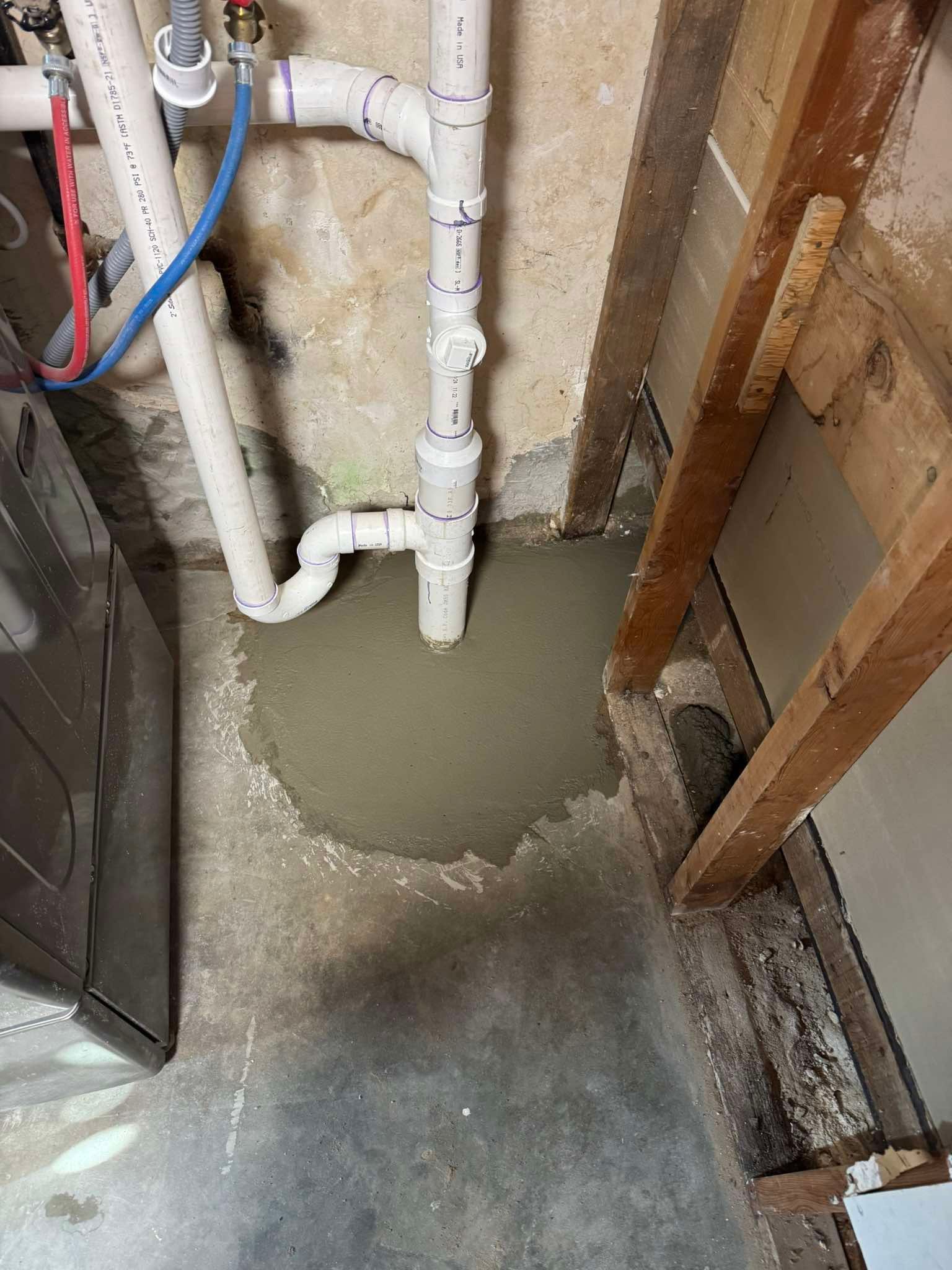 A vertical white PVC pipe enters a fresh patch of wet gray concrete on a basement floor next to unfinished wood framing.