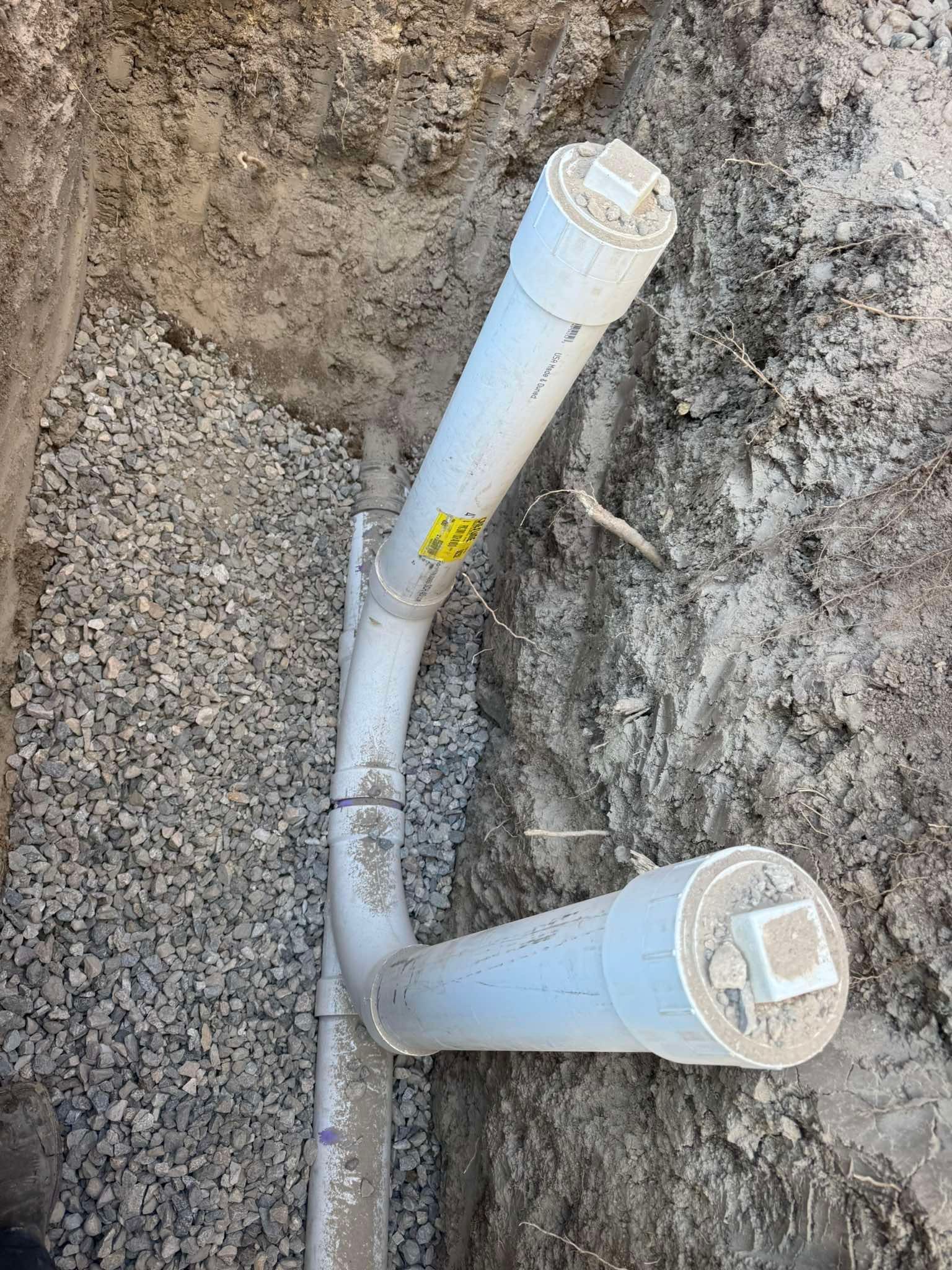 A white PVC pipe assembly with two capped access points, set in a gravel-lined dirt trench for underground plumbing.