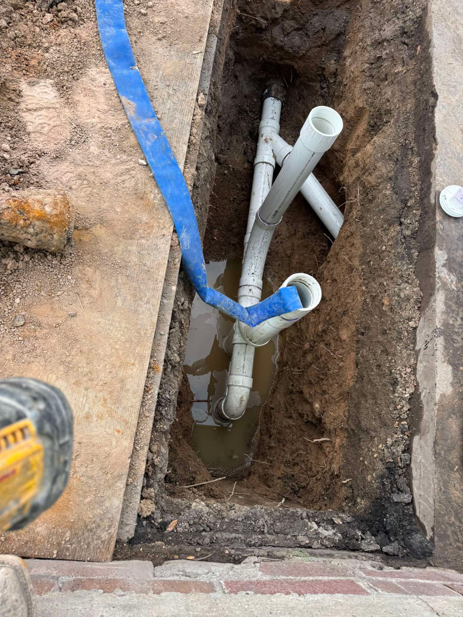 White PVC plumbing pipes emerge from a muddy trench in the ground, with a blue hose resting across them.