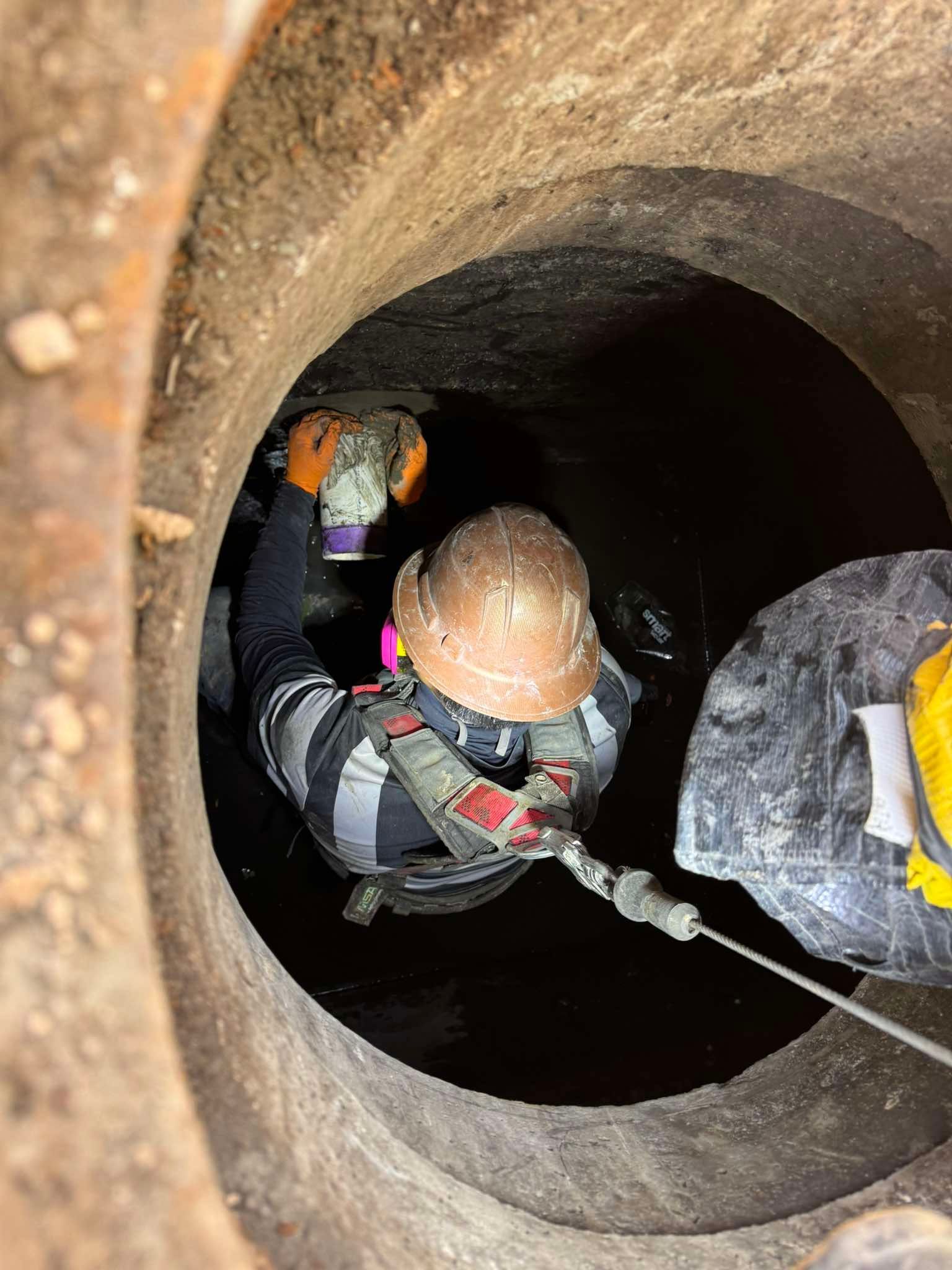 A worker in a helmet and harness is inside a manhole, performing maintenance.