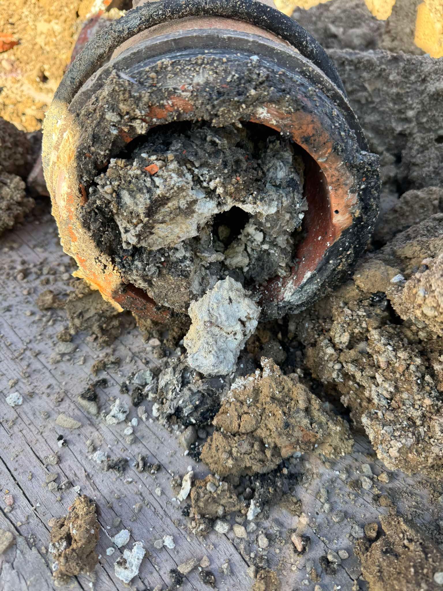 A close-up view of a broken, mud-caked pipe section filled with compacted debris and dirt, resting on wooden boards.