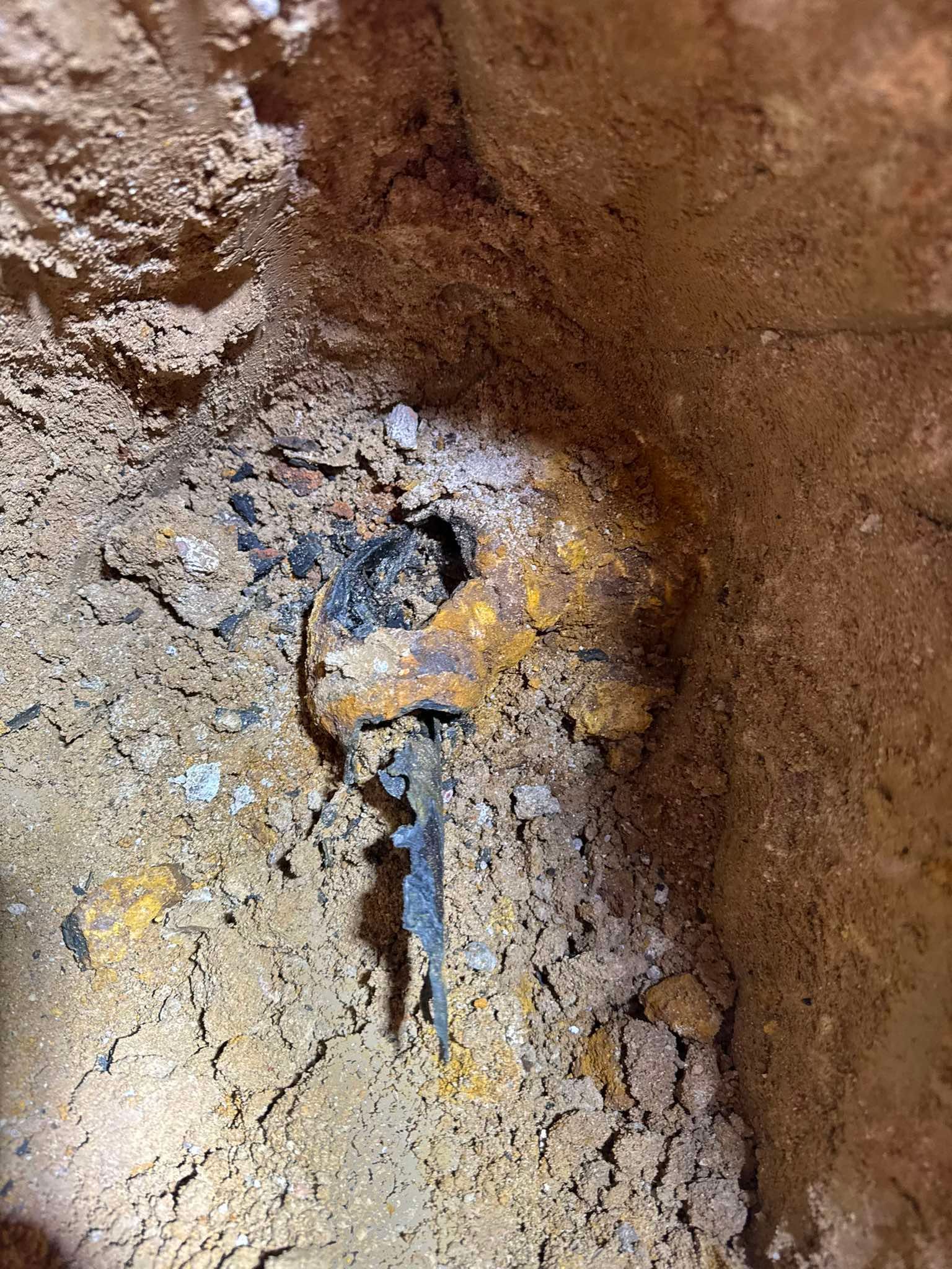 An excavated hole in reddish soil containing a corroded, rusted metal object with a jagged edge.