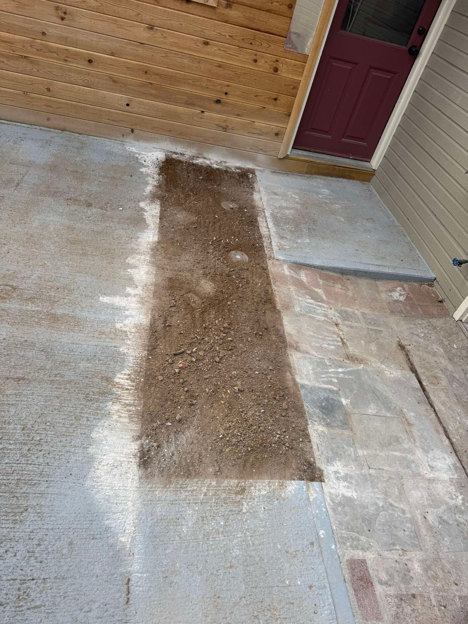 A rectangular dirt patch fills a gap between concrete patio sections near a house with wood siding and a burgundy door.