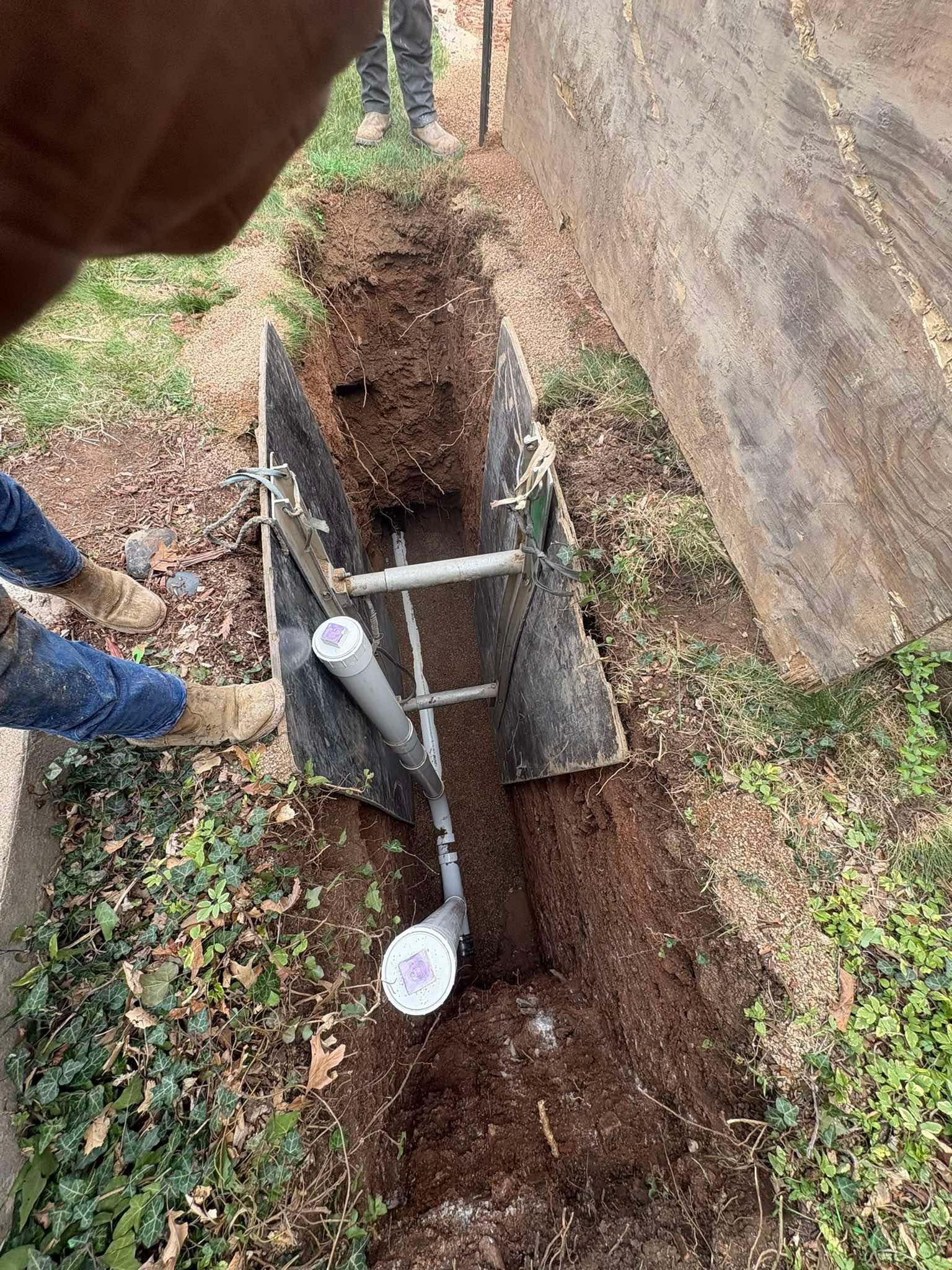 A construction trench containing grey PVC piping and metal shoring panels in a dirt yard next to a building wall.