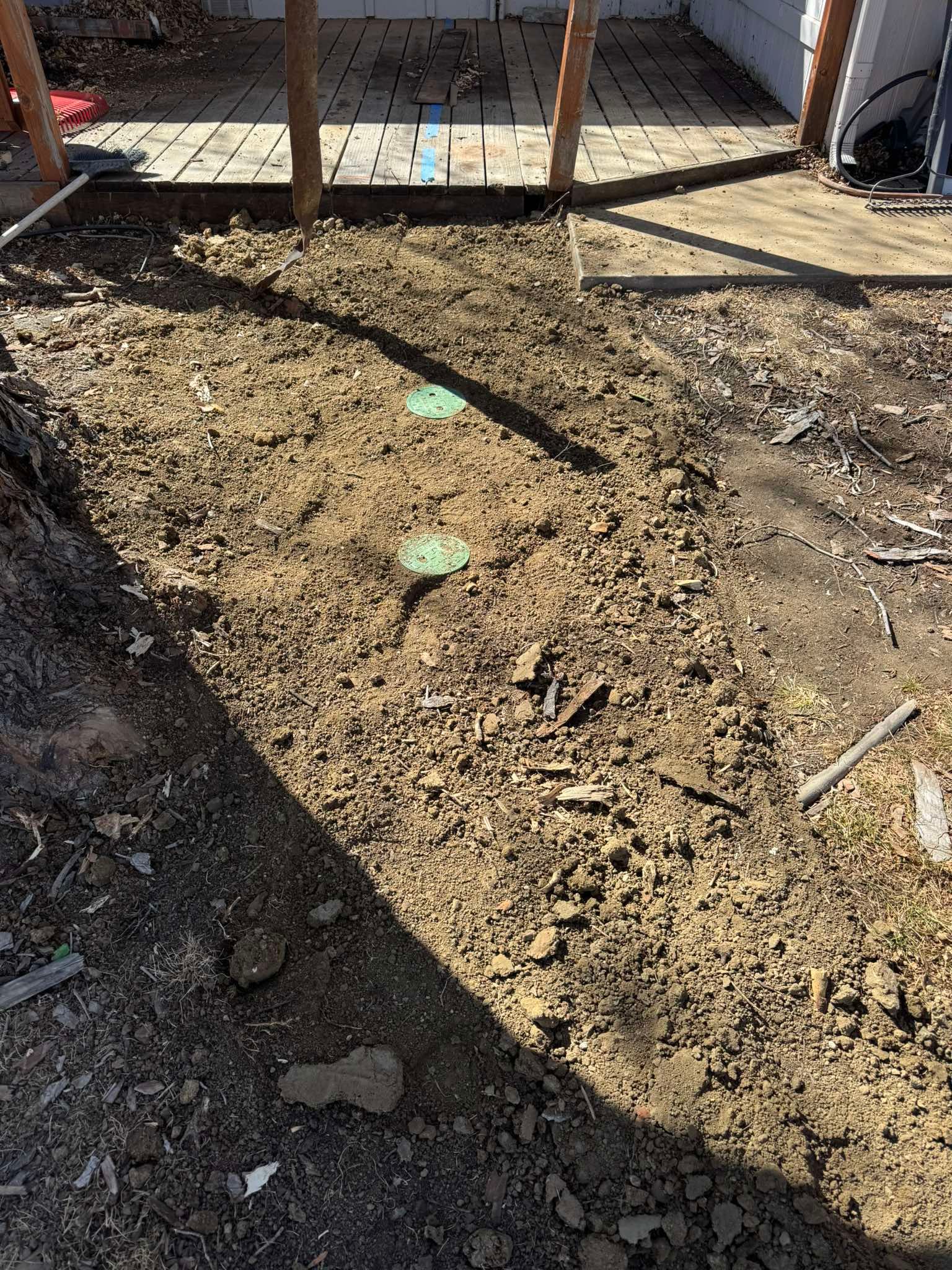 Two circular green utility access covers are embedded in a strip of bare dirt in a backyard near a wooden deck.