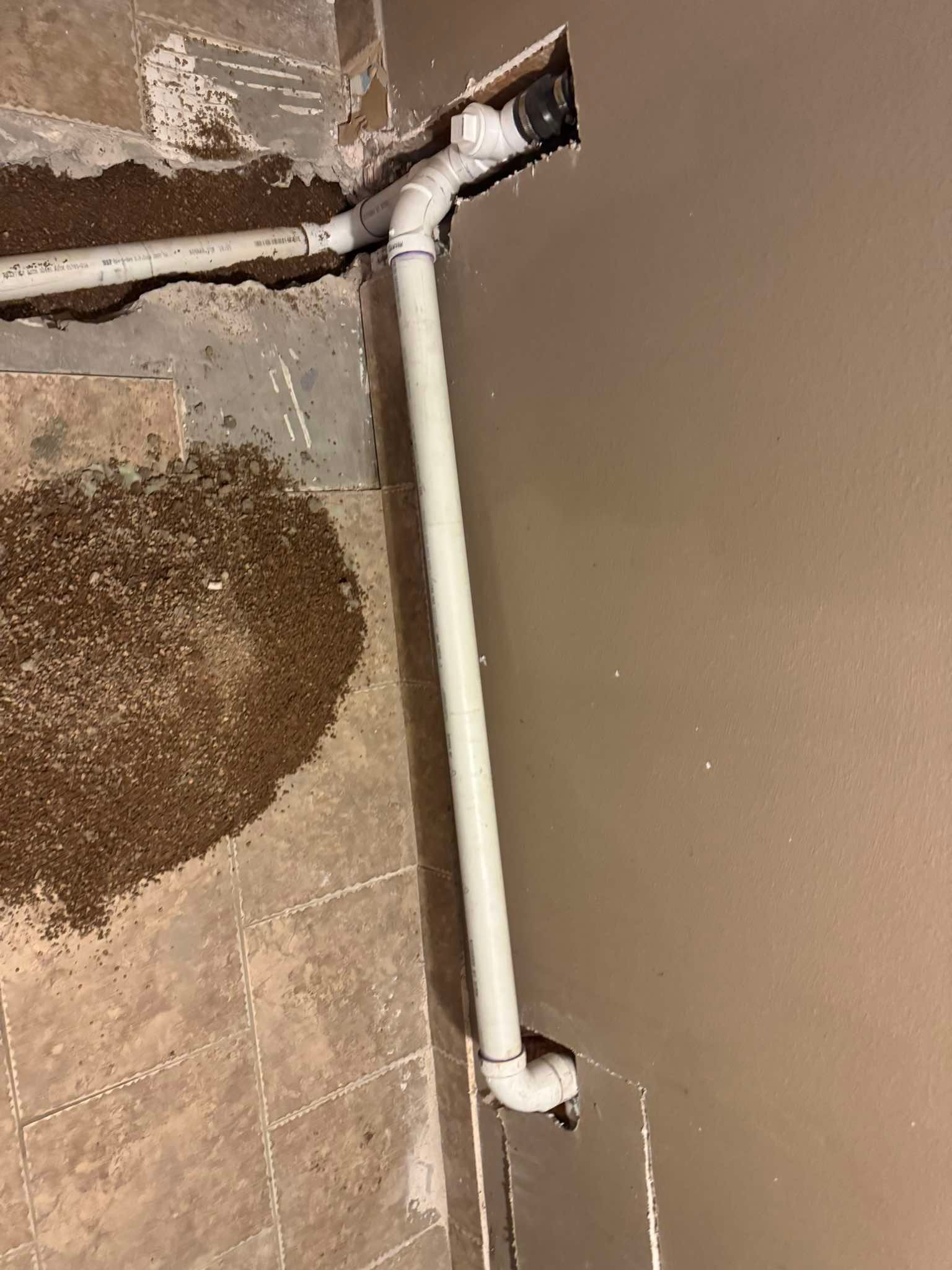 White PVC pipes connected by a Y-fitting, running along a wall and across a floor with scattered debris and grout lines.