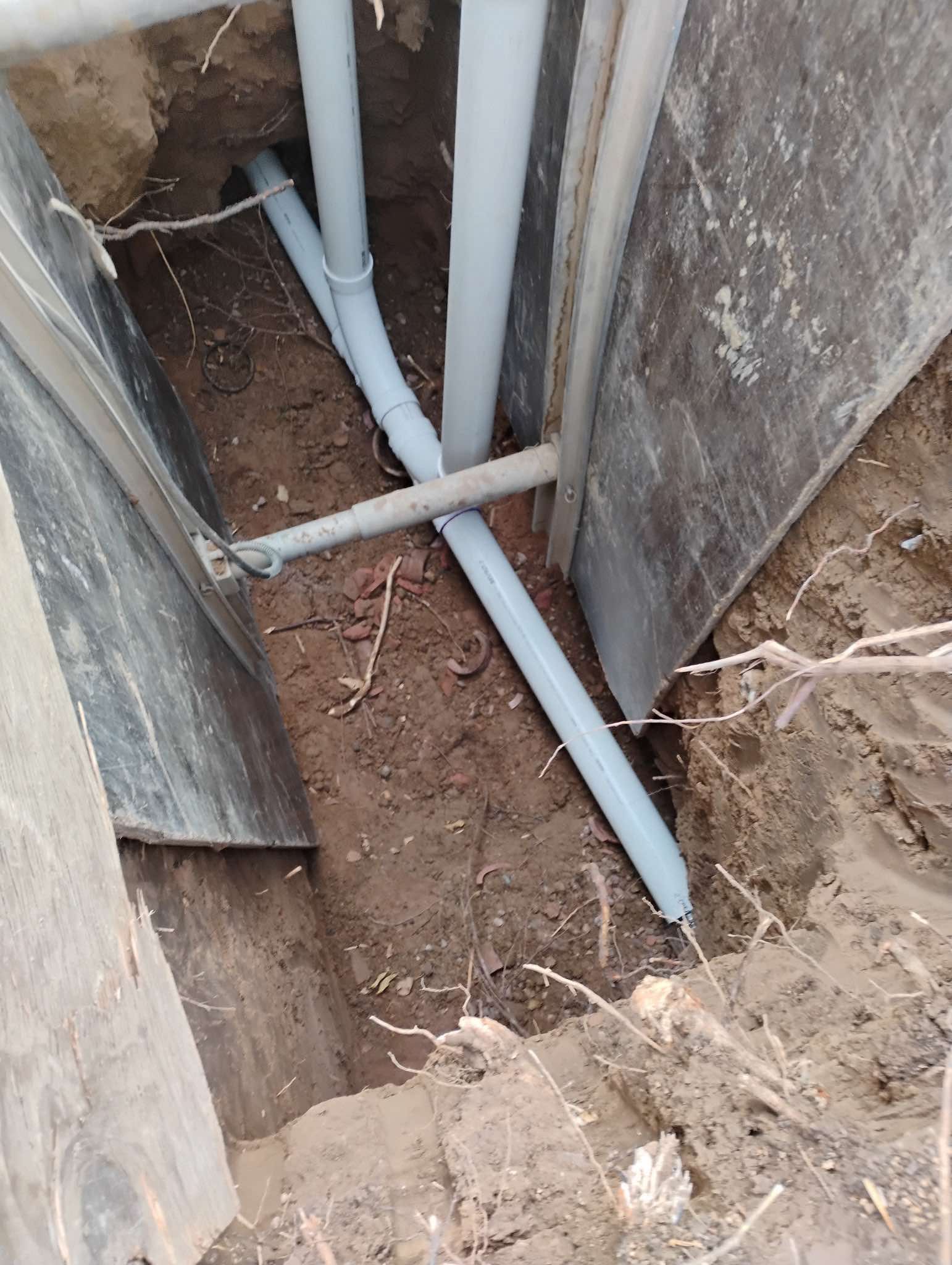 A high-angle view of gray PVC pipes laid in a narrow dirt trench between wooden shoring boards.