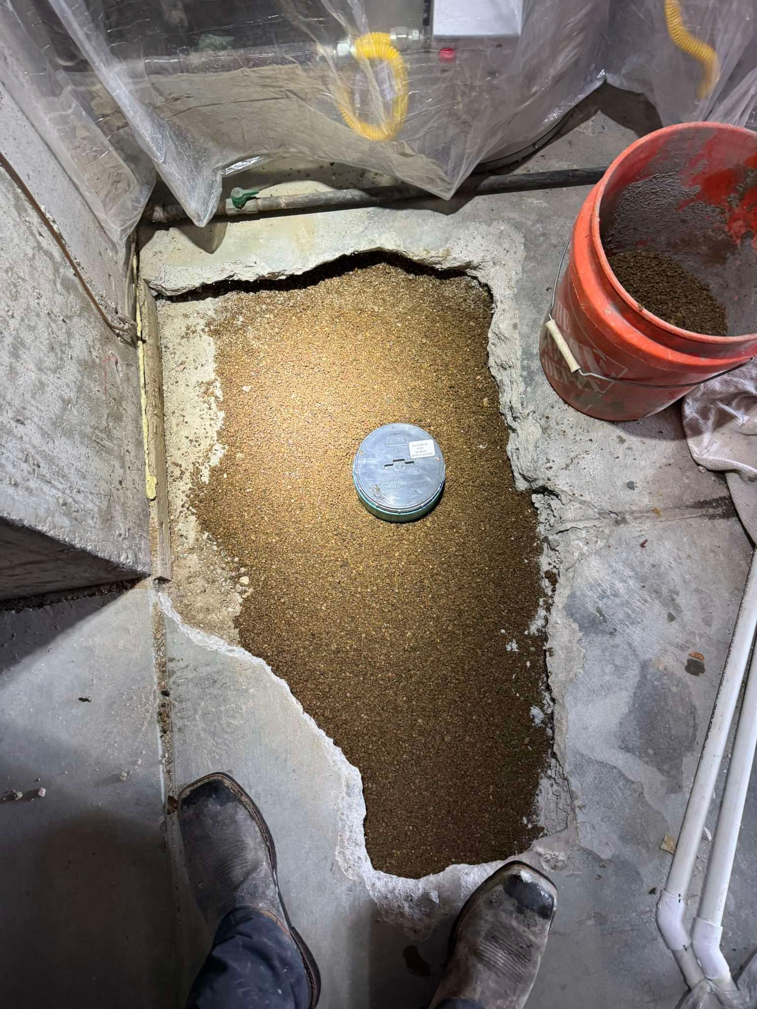 A person stands over a patch of exposed gravel in a concrete floor, with a circular plumbing access cover in the center.
