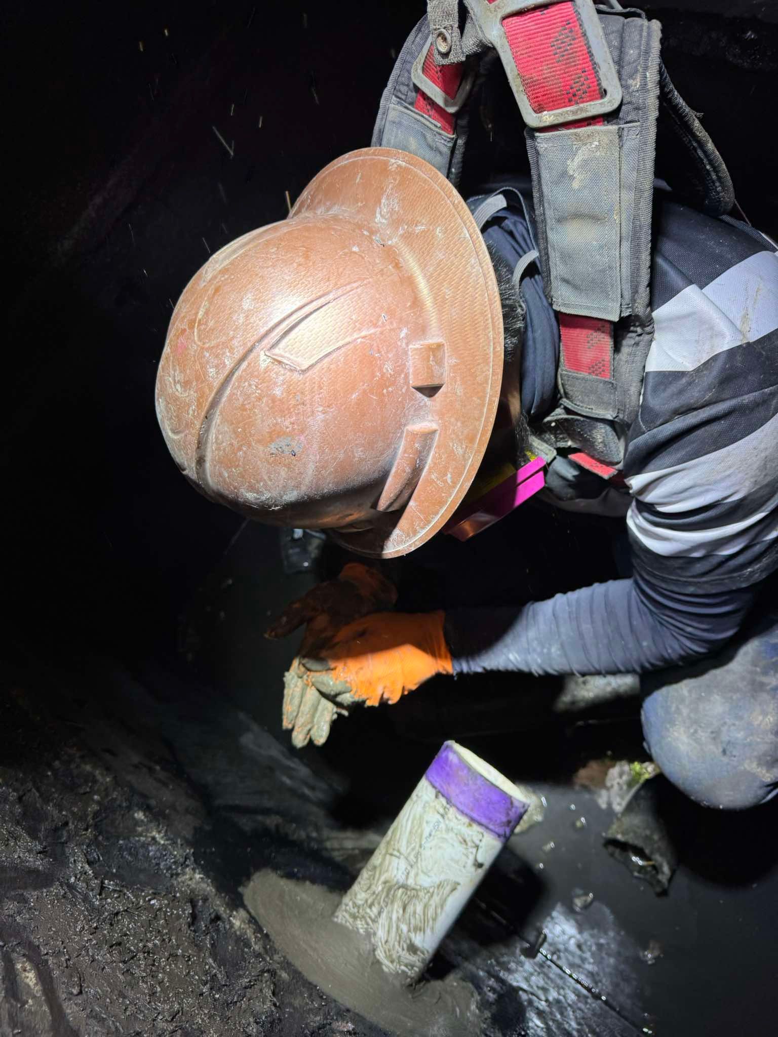 A worker in a hard hat and safety harness crouches in a dark, muddy space, holding a pipe section with orange gloves.