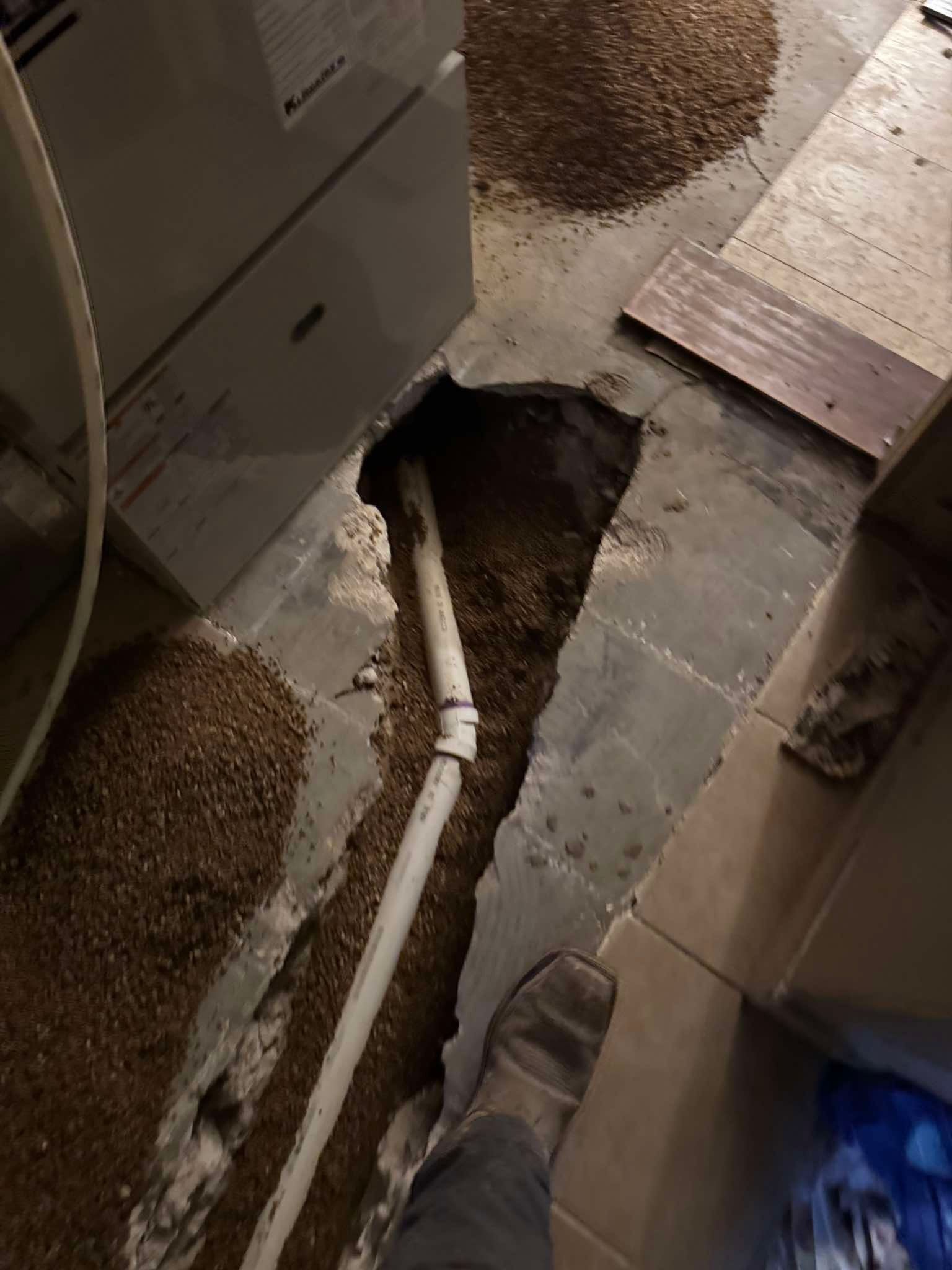 A trench cut through a concrete basement floor, exposing dirt and a section of white PVC piping near a furnace.
