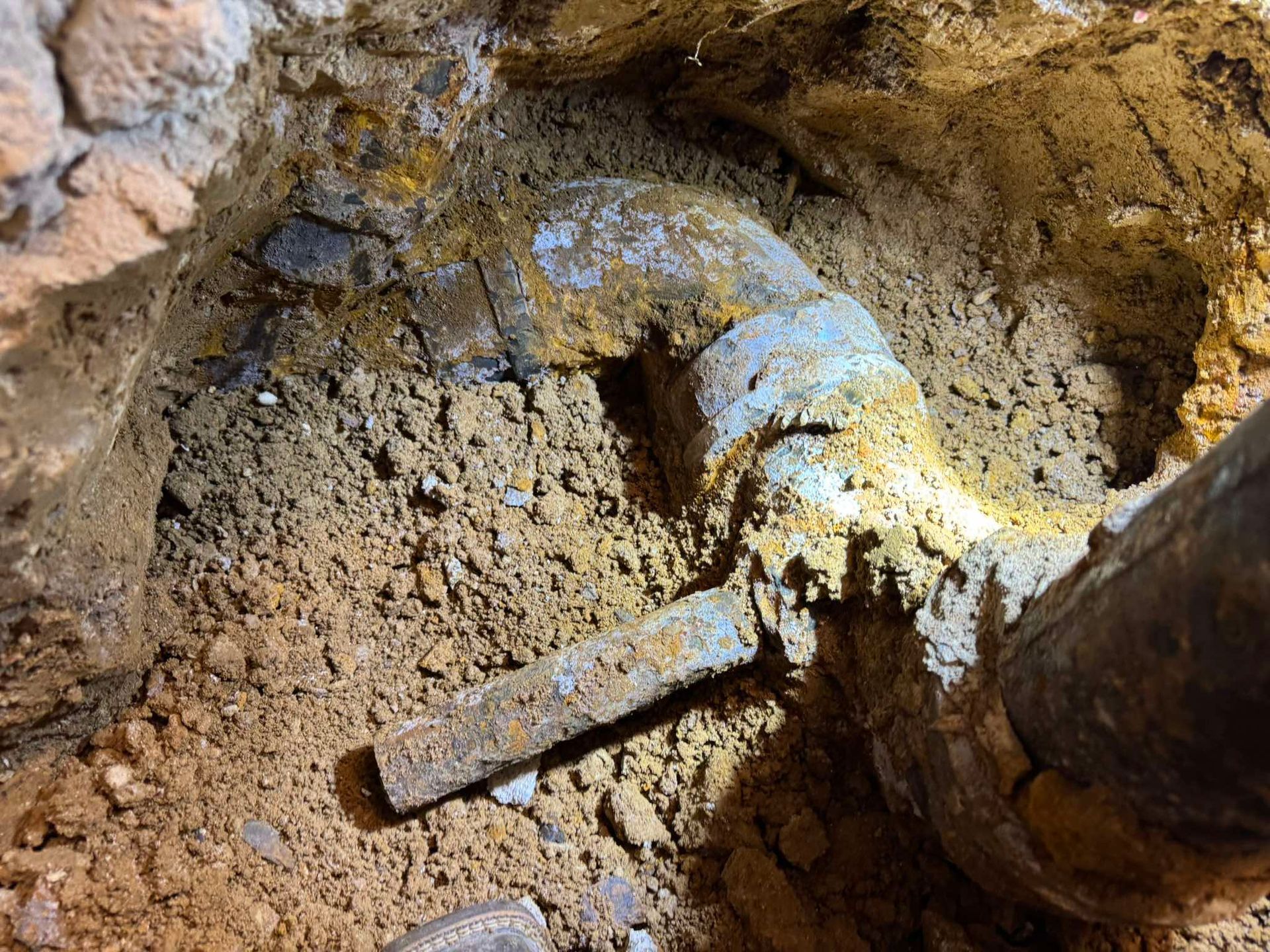 A close-up view of an exposed, aged metal plumbing pipe joint buried in dirt during an excavation or repair.