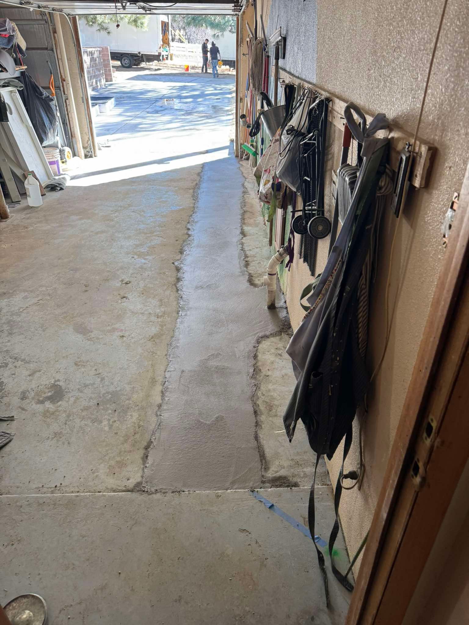 A concrete patch on a garage floor leads toward an open door, with tools hanging on a wooden wall to the right.