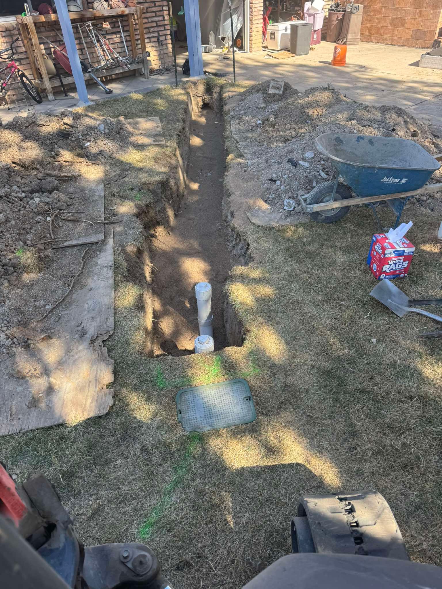 A dug trench in a backyard with a white PVC pipe rising from the ground, a wheelbarrow nearby, and scattered dirt.