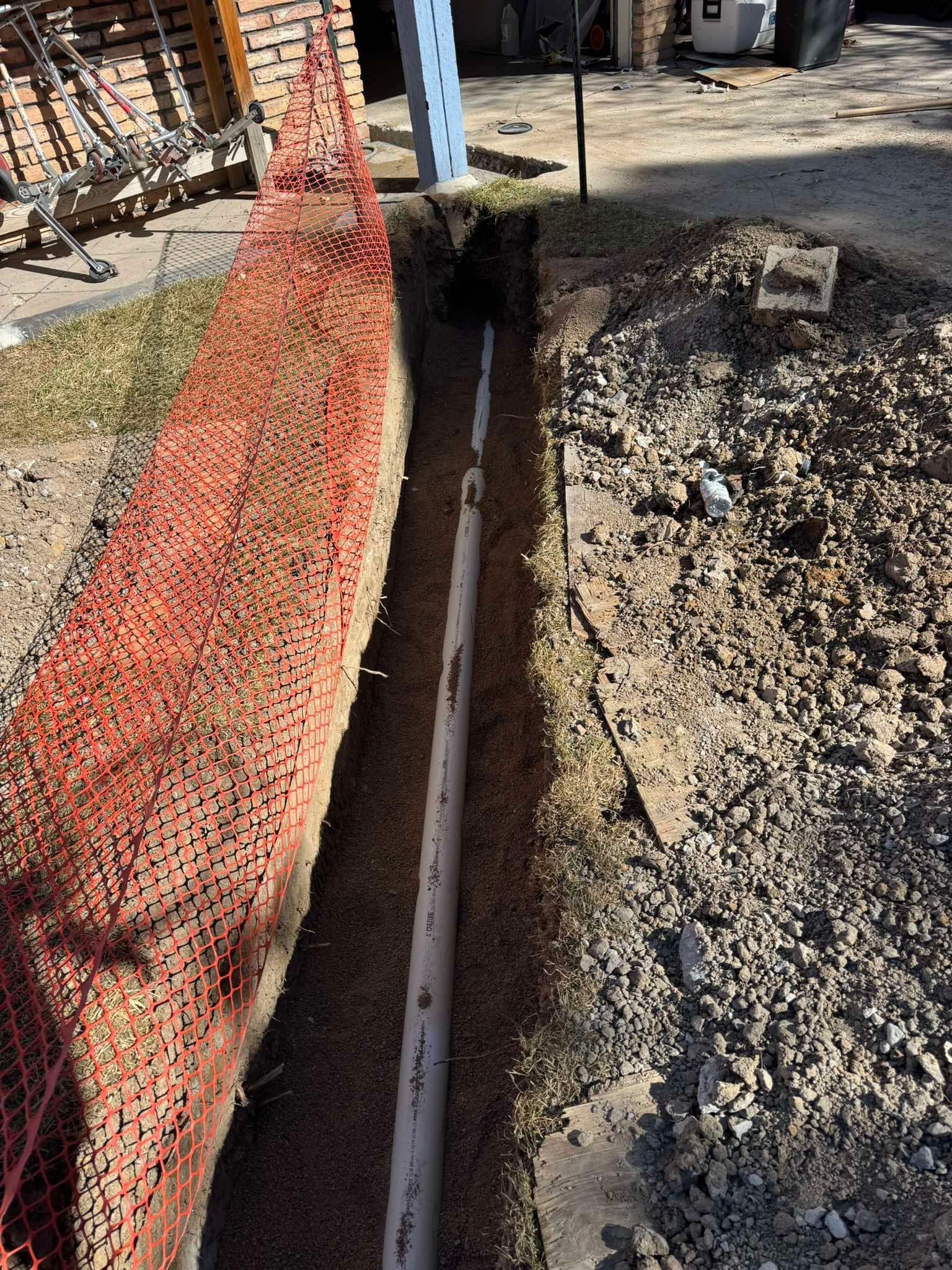A white plastic pipe sits at the bottom of a narrow construction trench next to orange safety fencing.