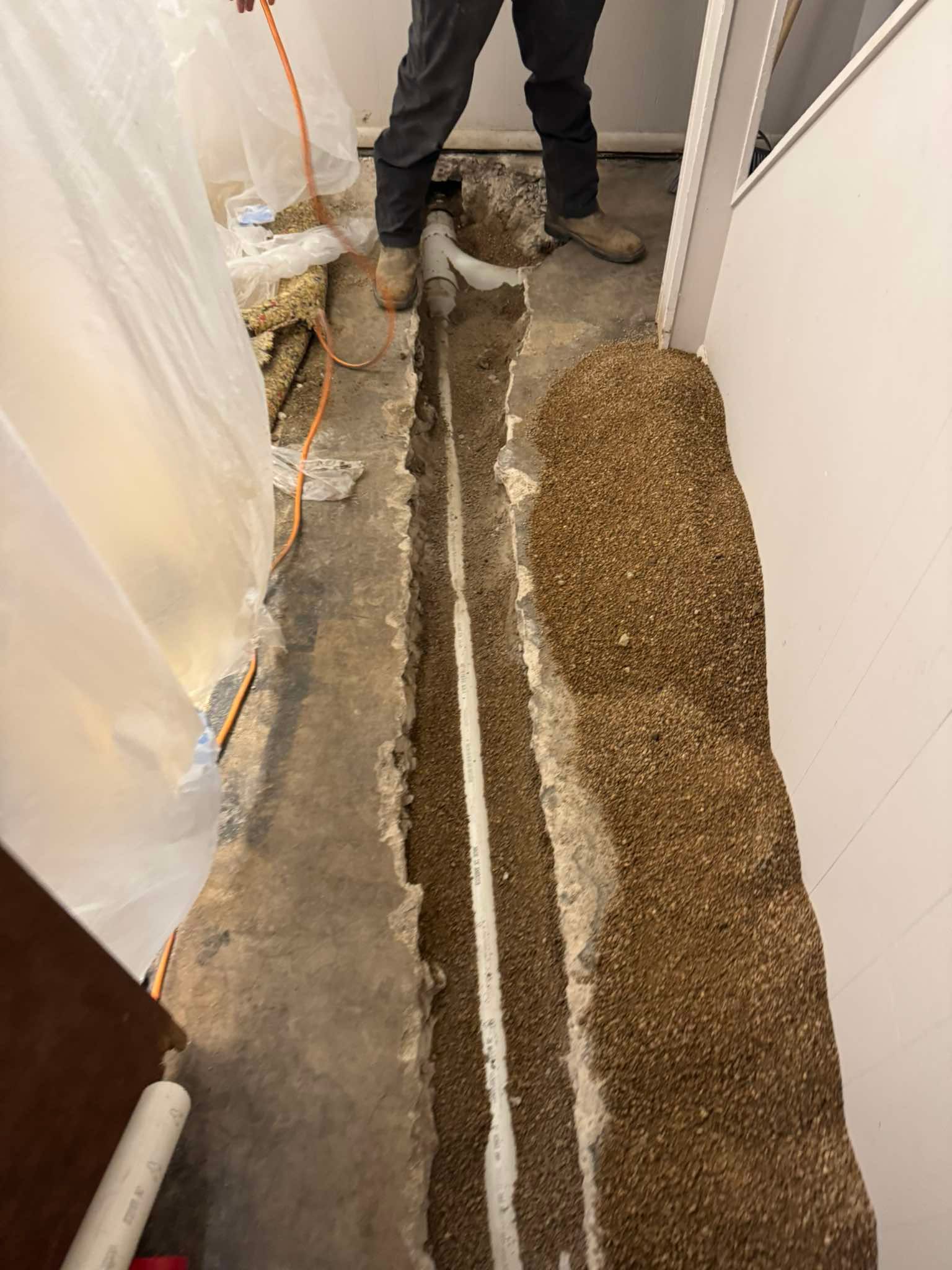 A trench cut into a concrete floor, revealing a white PVC pipe bedded in gravel, with a person standing nearby.