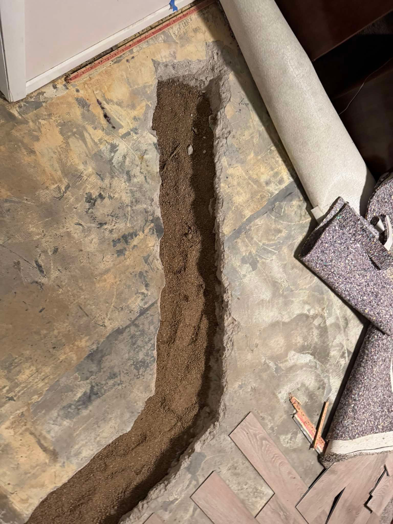 A trench cut into a concrete floor, revealing dirt and debris, with rolled-up carpet and flooring material nearby.