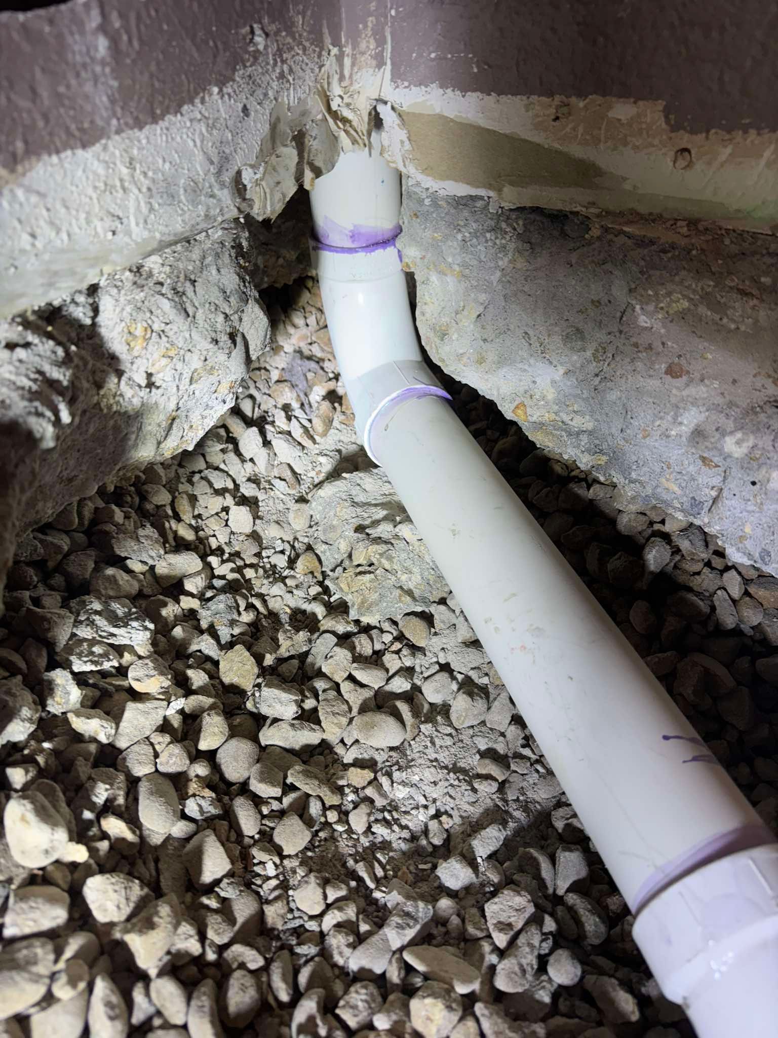 A white PVC drain pipe with purple-primed joints curves into a dirt and gravel trench near a concrete wall foundation.
