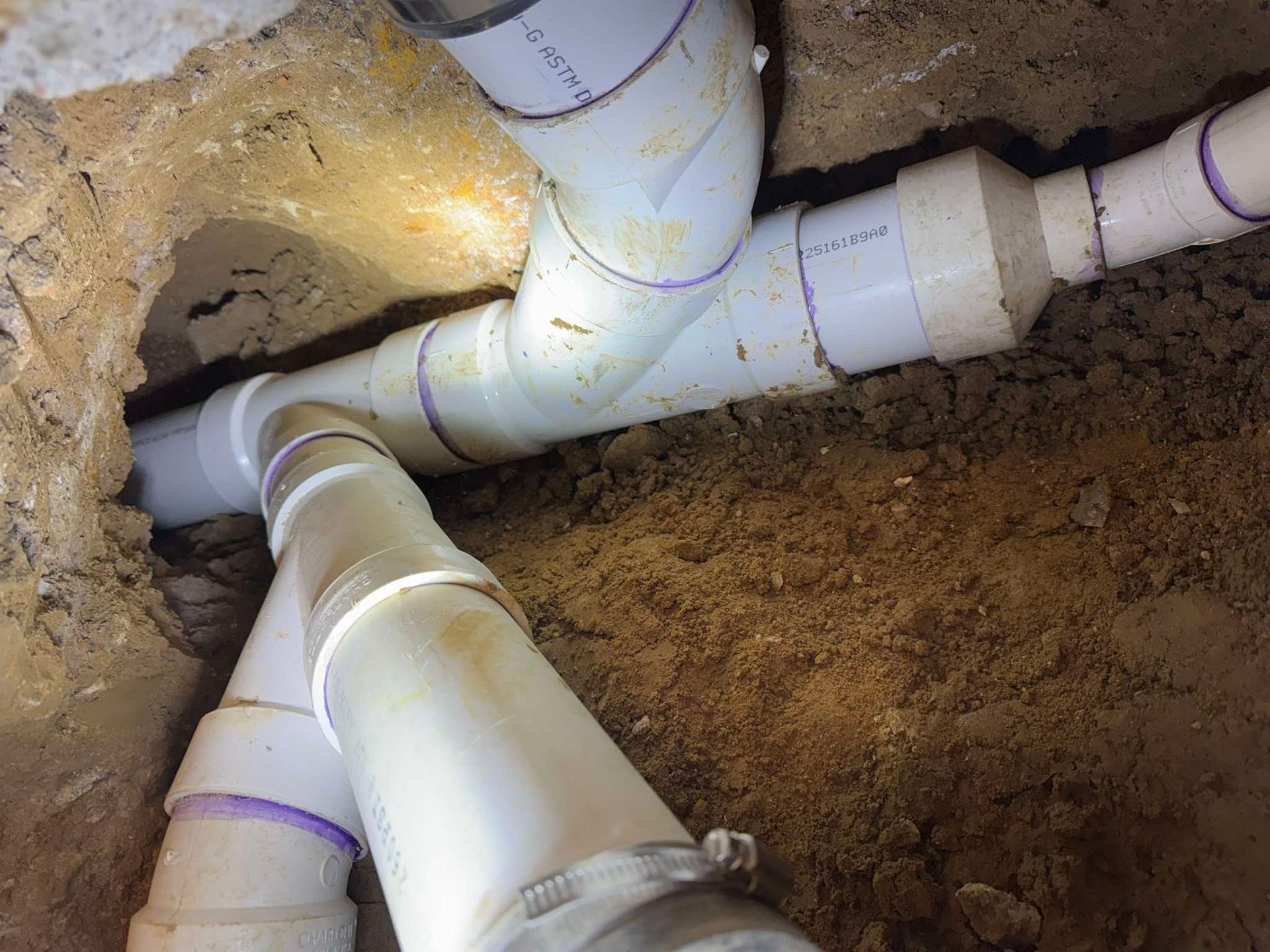 A close-up of white PVC plumbing pipes connected with white fittings, embedded in a hole dug into the dirt.