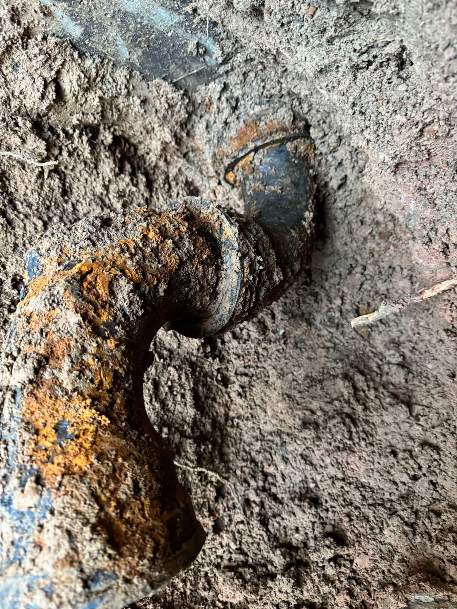 A heavily corroded, L-shaped cast iron pipe joint buried in moist, dark soil.