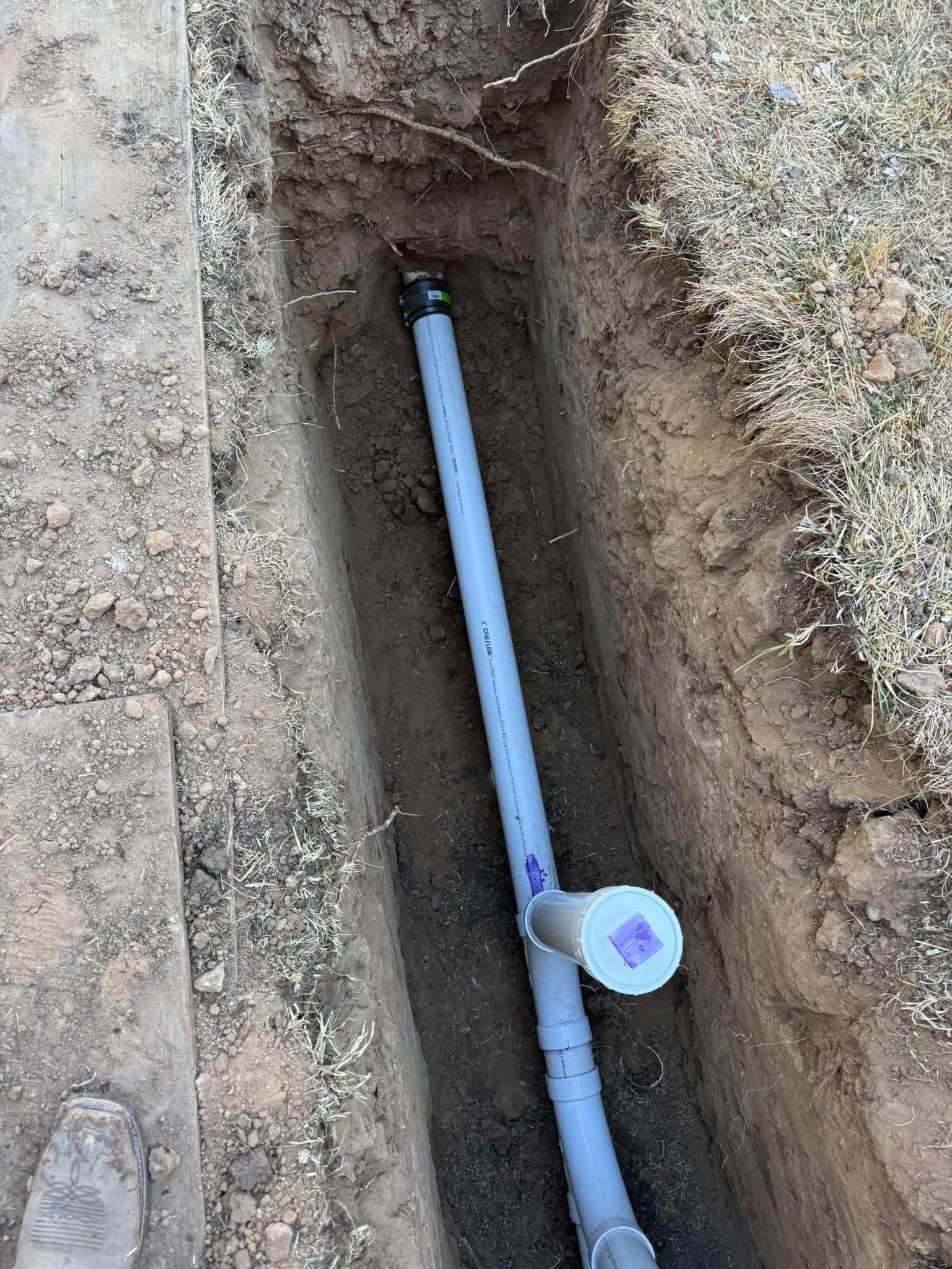 A grey PVC pipe with a capped vertical riser sits inside a narrow dirt trench.