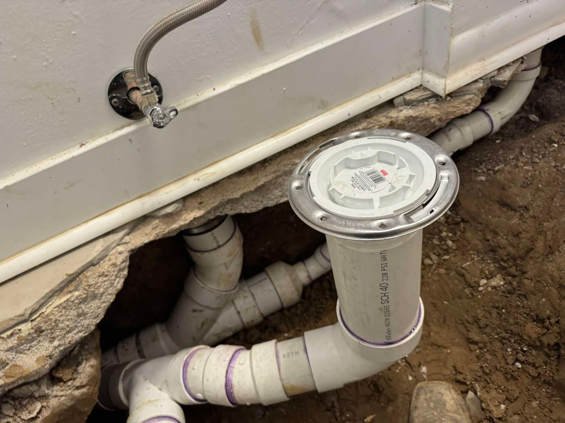 A white PVC toilet flange and drain pipe installed in a concrete floor trench next to a water supply line.