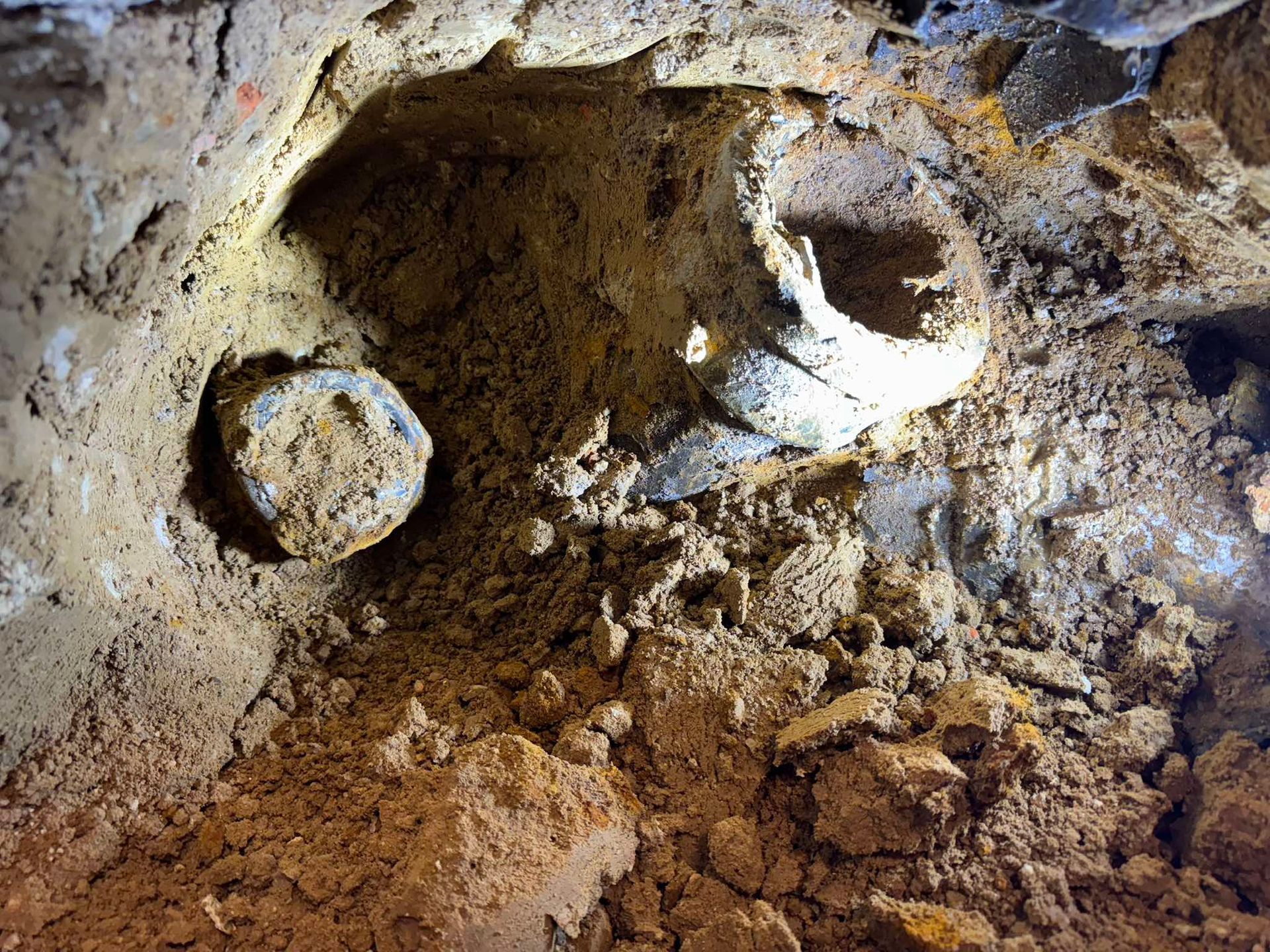 A hole excavated in soil revealing a section of dark, weathered, circular pipe embedded in the earth.