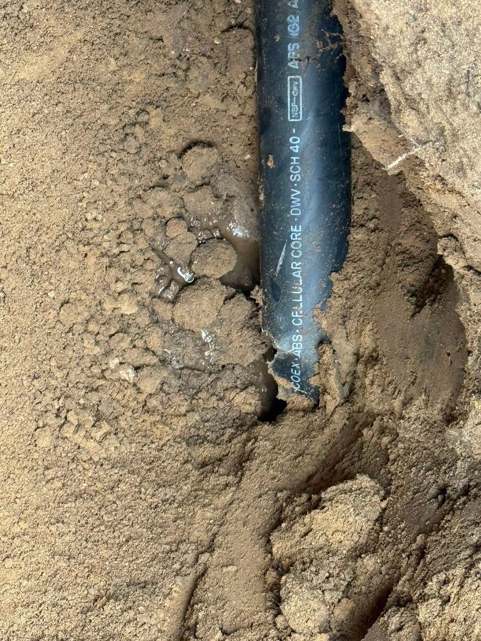 A black plastic pipe lies exposed in damp, sandy soil, showing signs of water leakage near the pipe's surface.