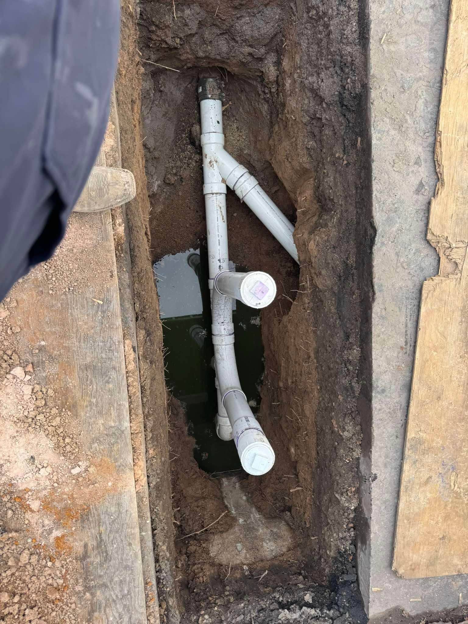 A trench in the ground reveals grey PVC plumbing pipes with capped ends and a Y-connector joint.
